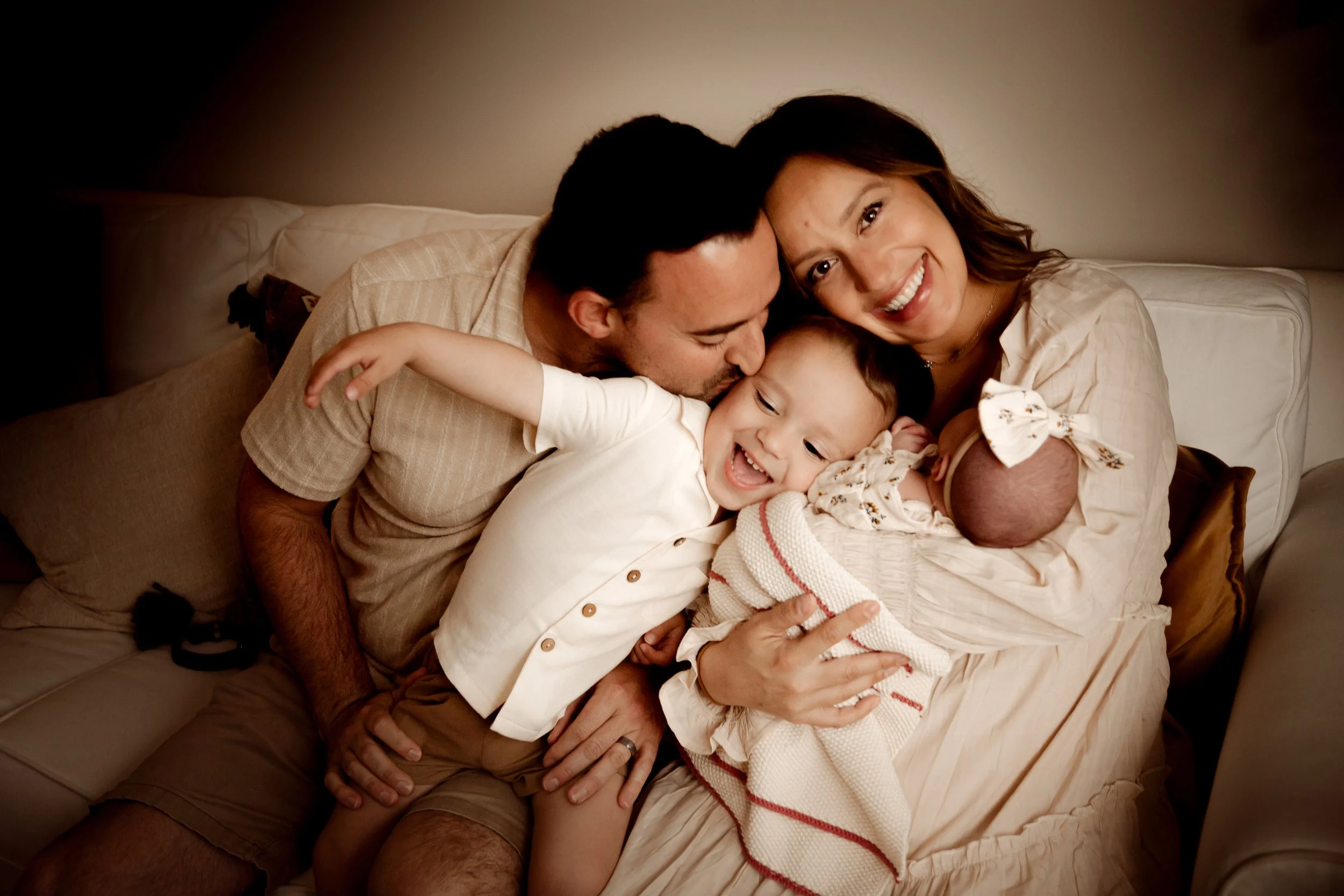 A happy family of four sitting on a beige couch, including a woman, a man, a young girl, and a baby, sharing a tender moment together.