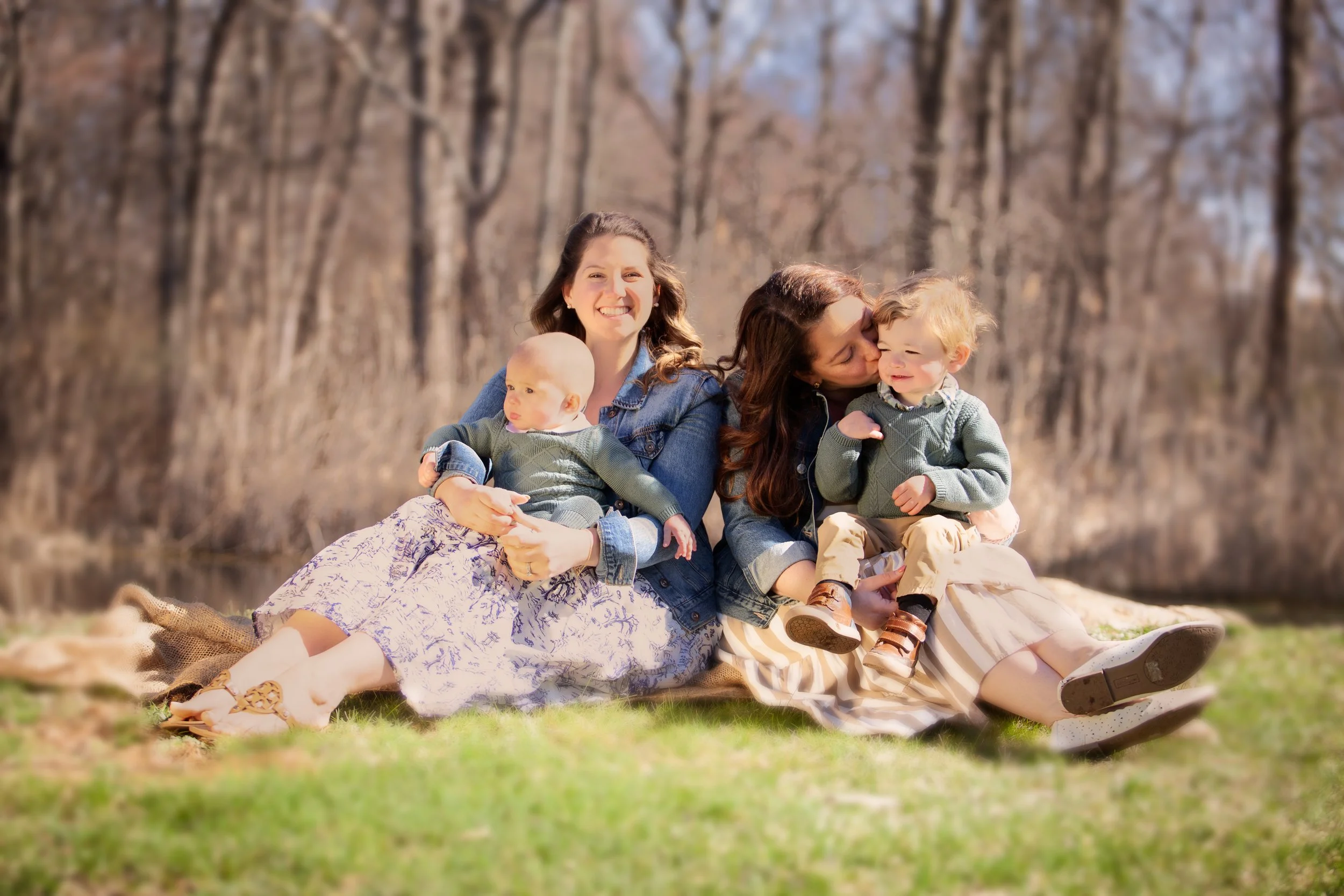 Two women and two children sitting on a blanket in a park with bare trees in the background. The woman on the right is kissing a young boy, and the woman on the left is holding a baby.