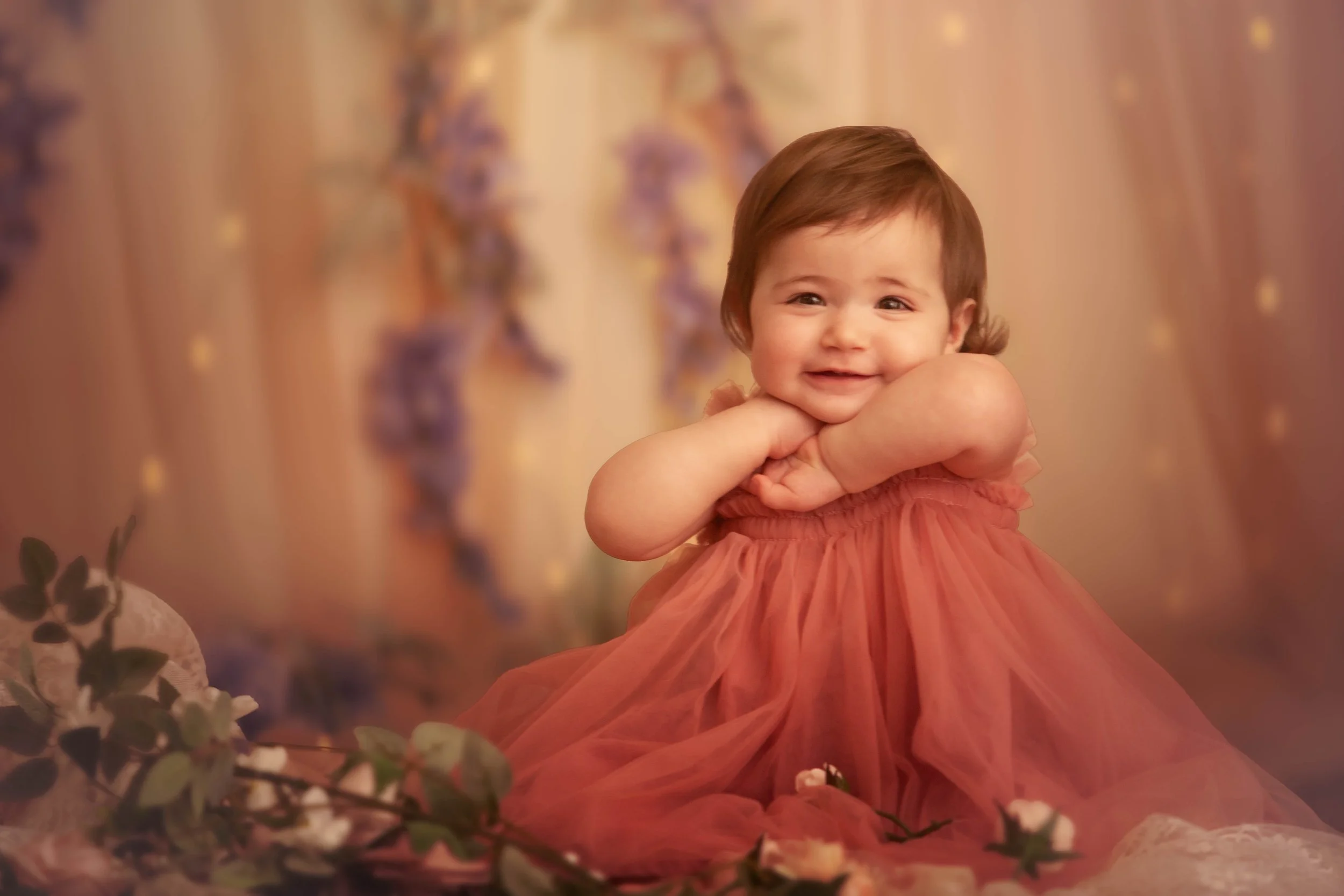 A smiling baby girl with short brown hair, wearing a flowing pink dress, sitting on a decorated surface with plants and flowers, with a blurred purple and yellow background.