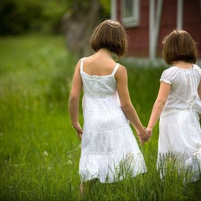 Two young girls in white dresses holding hands and walking through green grass outdoors.