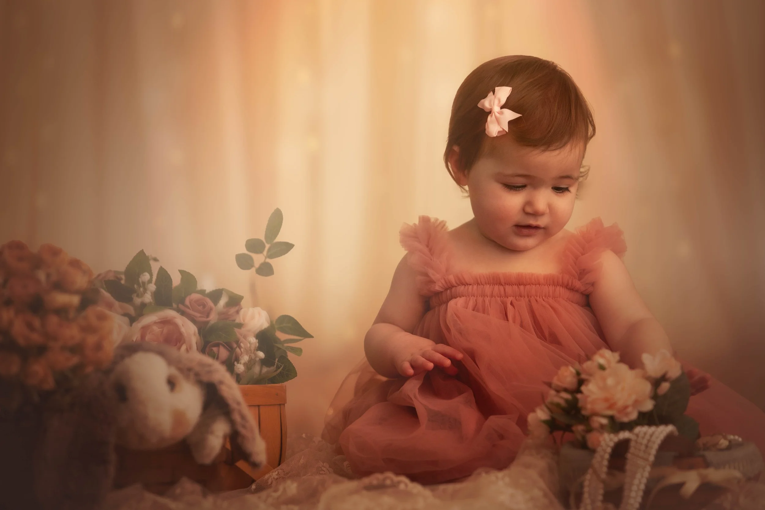 A young girl in a peach-colored dress sitting on the floor surrounded by flowers and plush toys, with warm lighting in the background.
