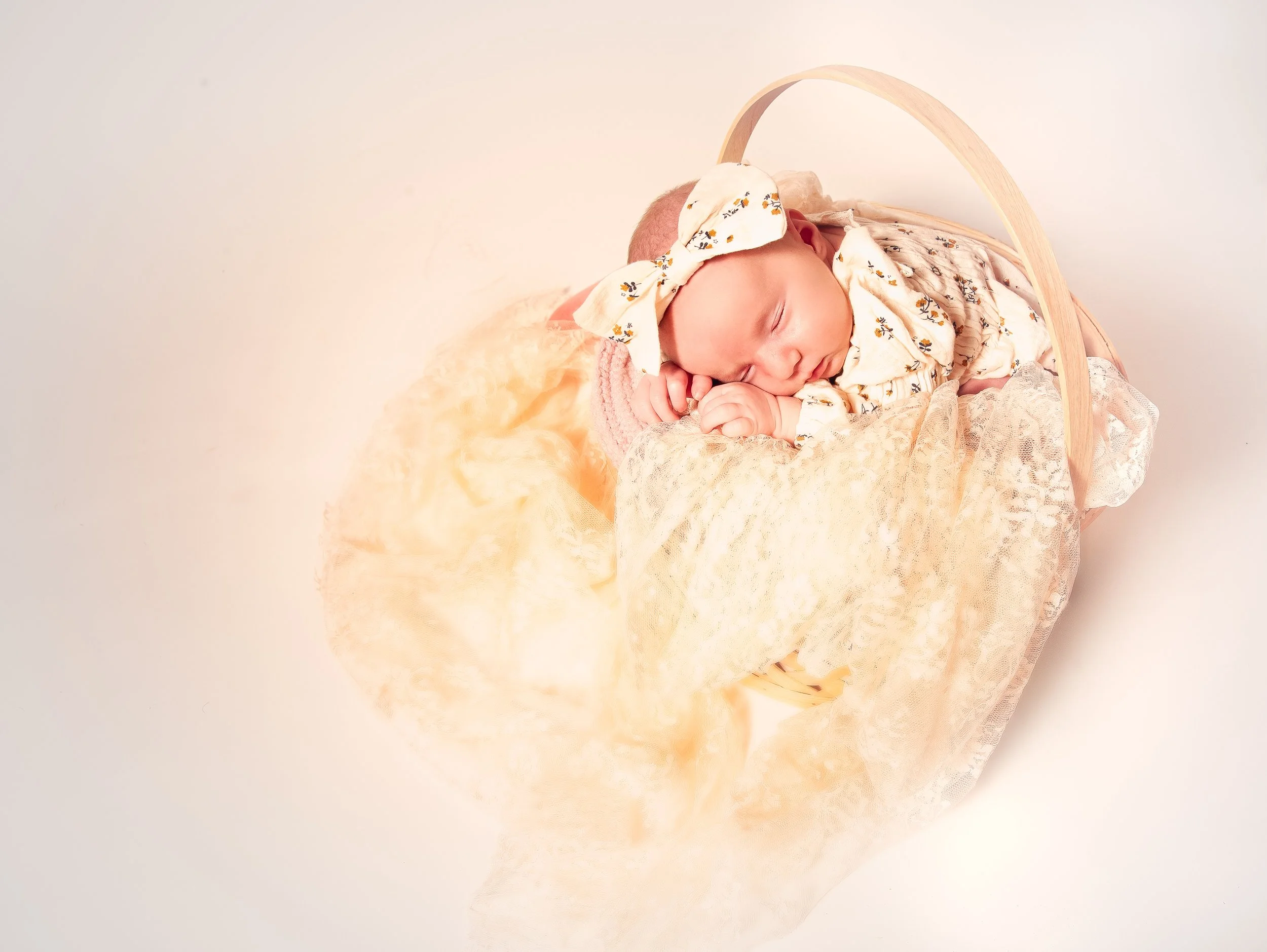 A sleeping baby with a headband, lying in a wicker basket decorated with lace and soft fabric.