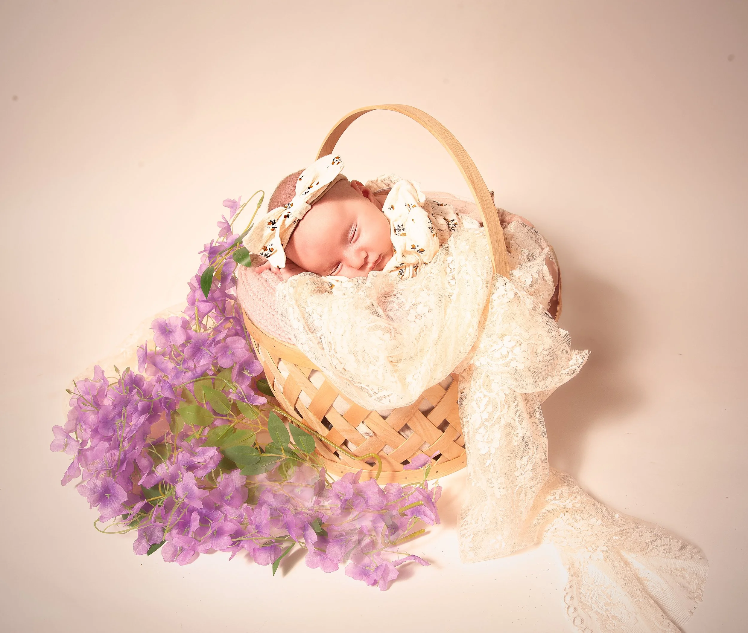 A newborn baby sleeping in a wooden basket decorated with pink flowers, dressed in lace and floral clothing, with a matching headband.