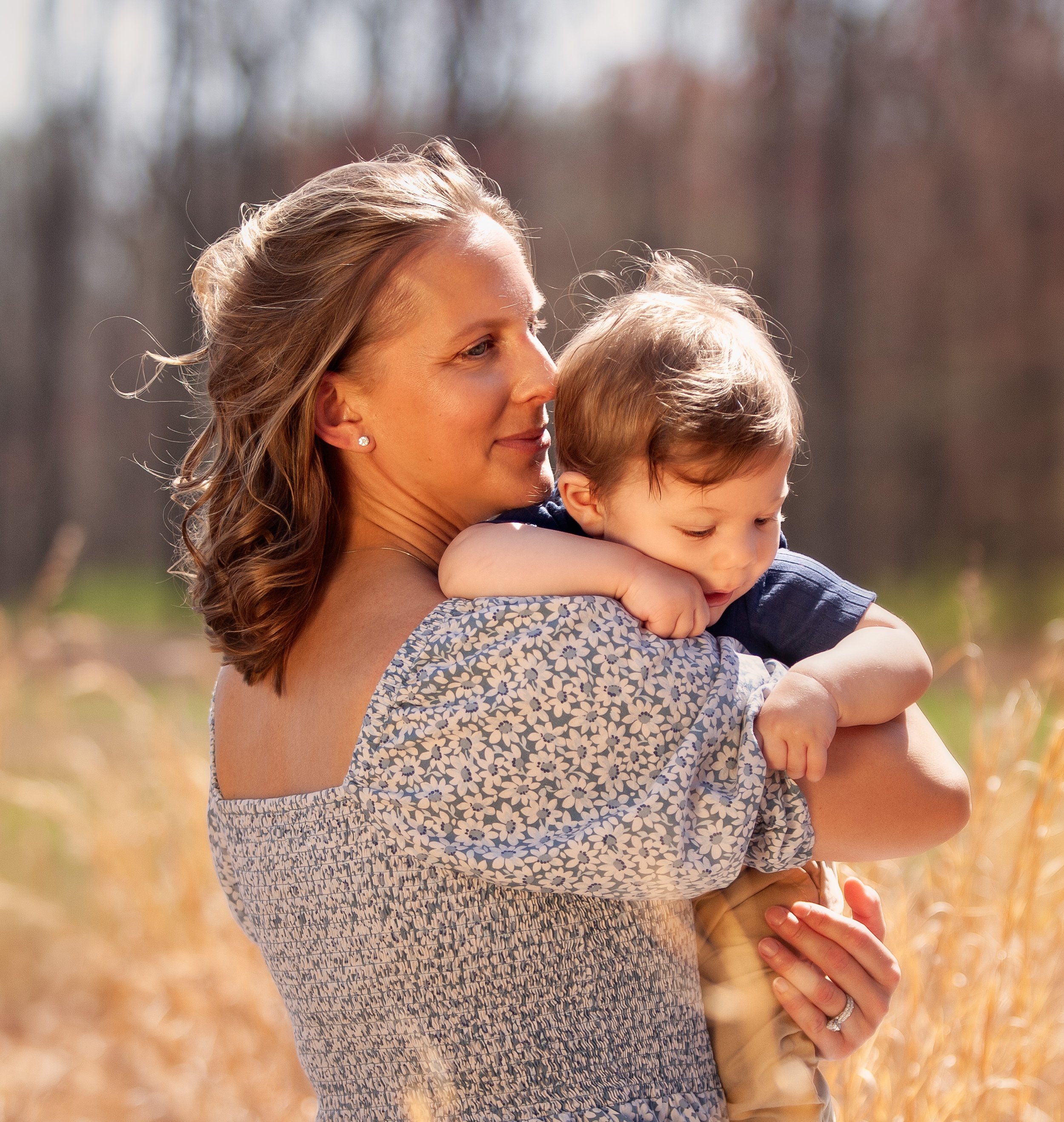 A woman holding a young boy in an outdoor setting during daylight. The woman's face is close to the boy's head, with a soft smile and blonde hair. The boy, with closed eyes, rests his head on her shoulder, and the background is blurred with trees and
