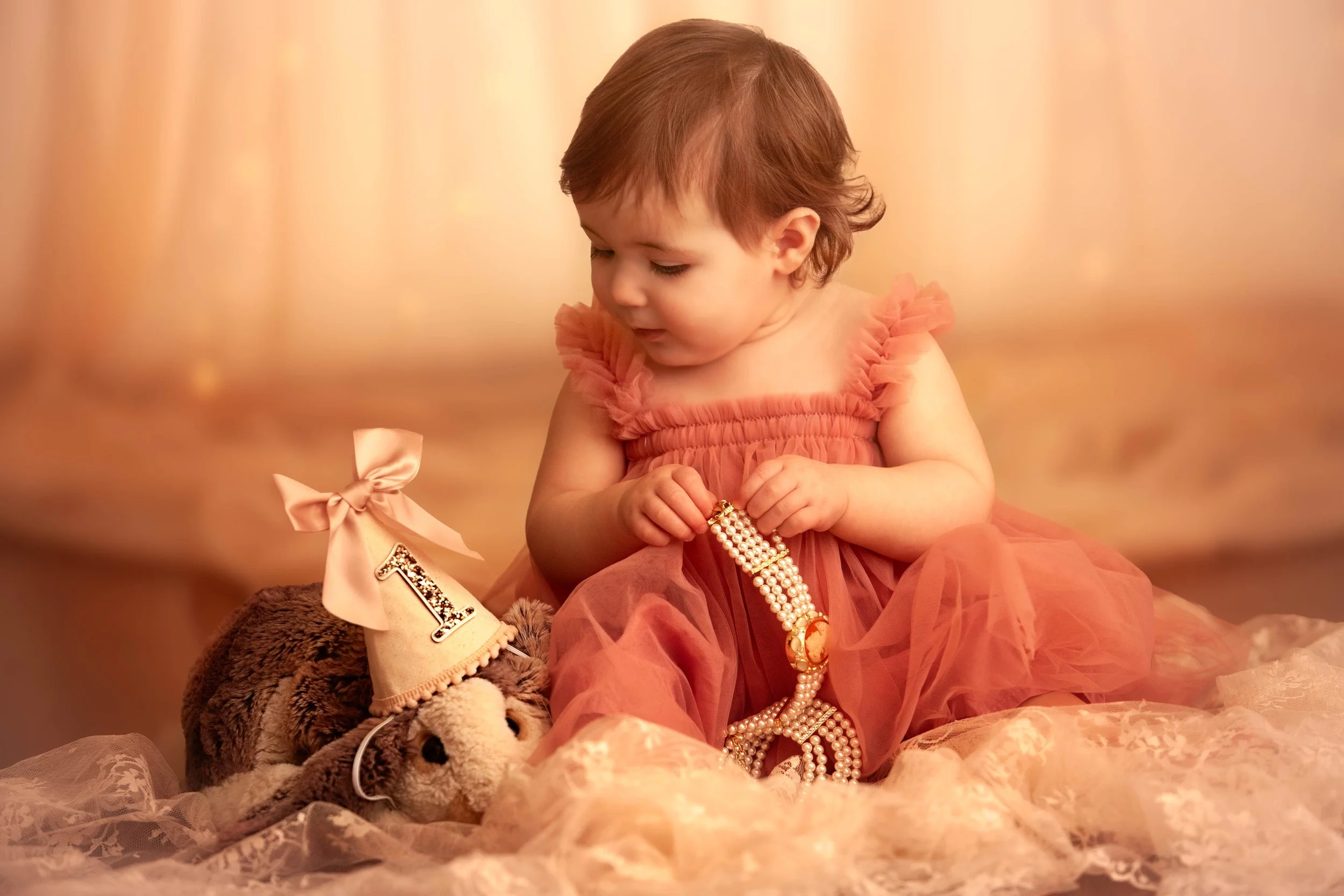 A young girl in a coral dress, sitting on fabric, holding a pearl necklace, with a stuffed animal wearing a party hat next to her.