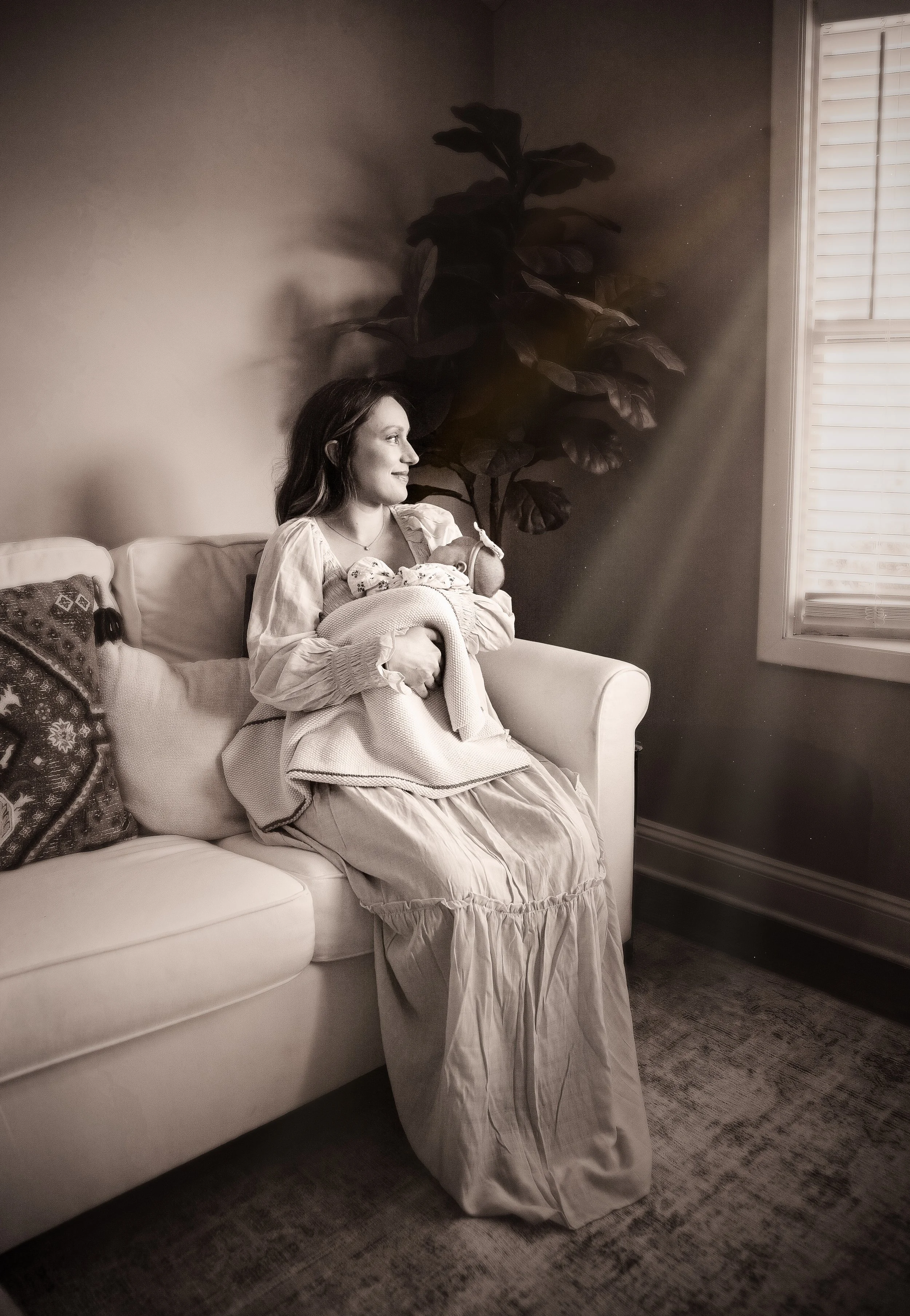 A woman sitting on a sofa holding a baby with a large houseplant behind her and a window with blinds.