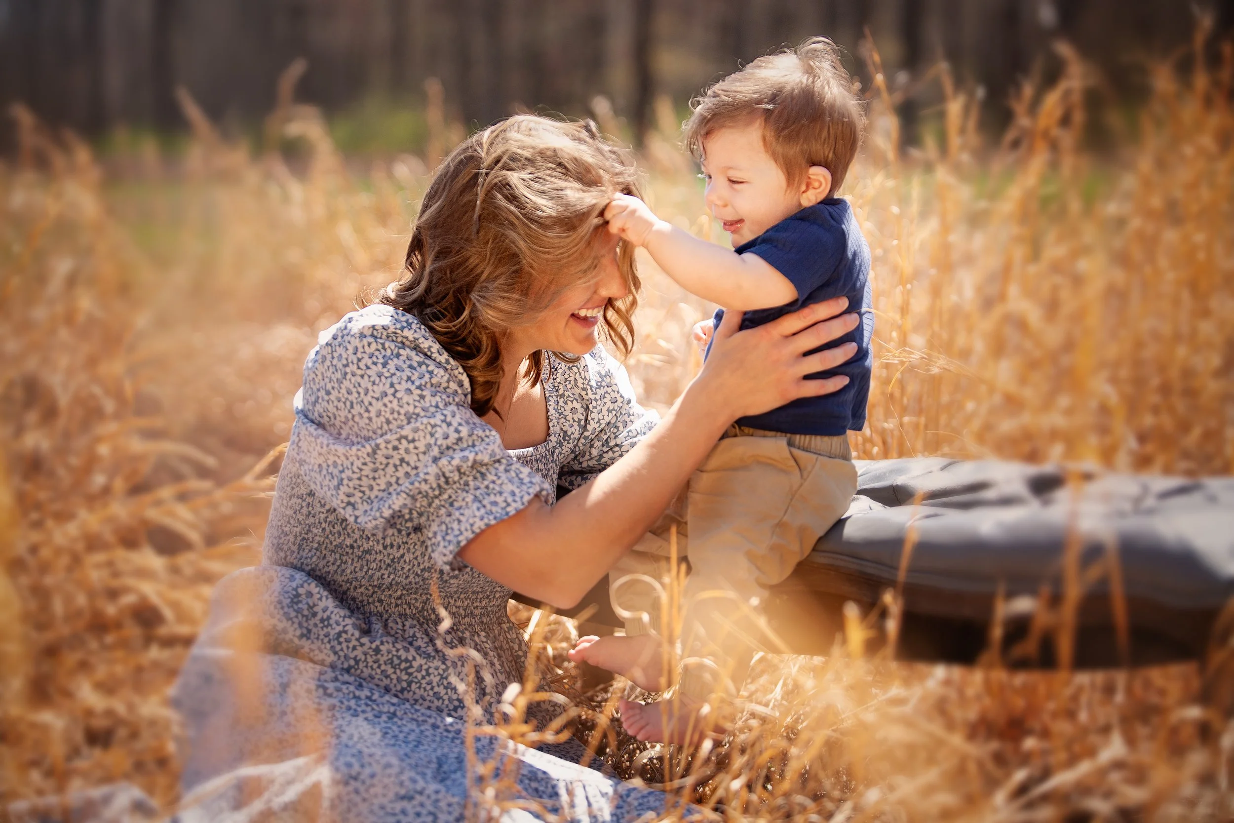A woman and a young boy outdoors in a field of tall, golden grass, smiling and playing together. The woman has curly hair and is wearing a patterned dress, while the boy has short hair and is wearing a dark blue shirt and tan shorts.