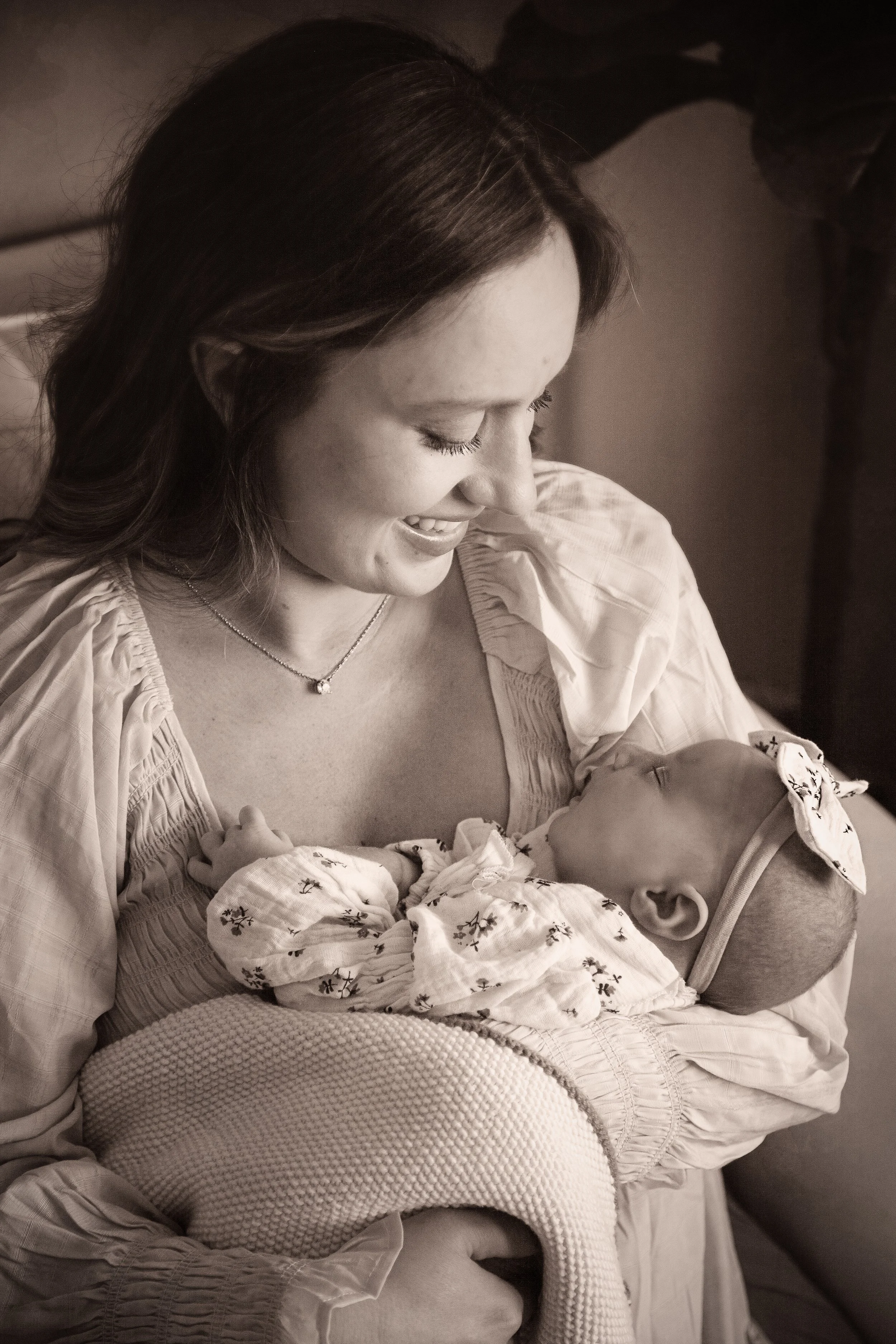 A woman with long dark hair wearing a light-colored blouse and a delicate necklace, smiling while holding a newborn baby in her arms. The baby is wrapped in a patterned blanket, and the woman appears to be looking lovingly at the infant.