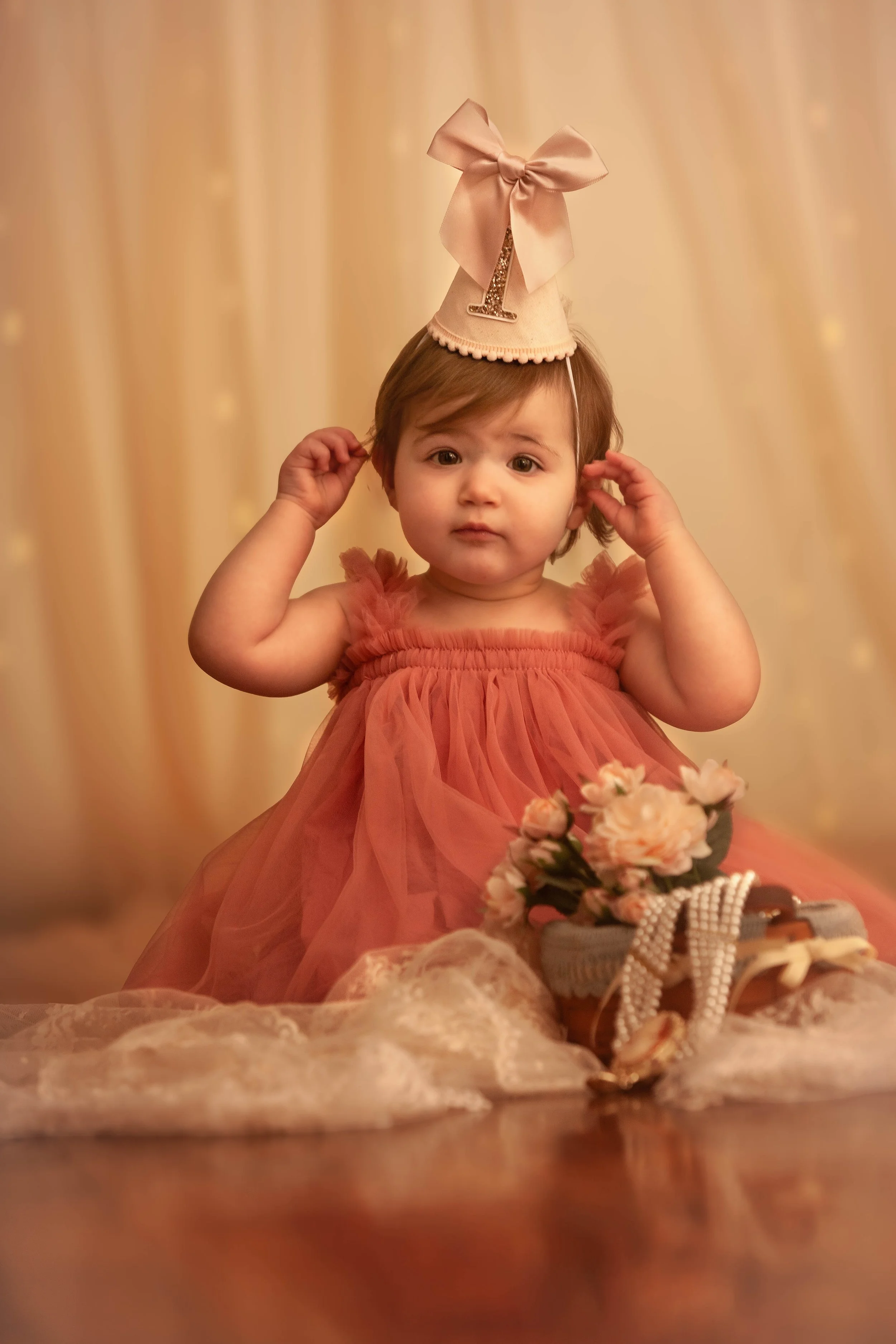 A young girl in a pink ruffled dress sits on the floor with a basket of pink flowers and pearls in front of her. She is wearing a birthday hat with a gold star and a large pink bow, and has a curious expression.