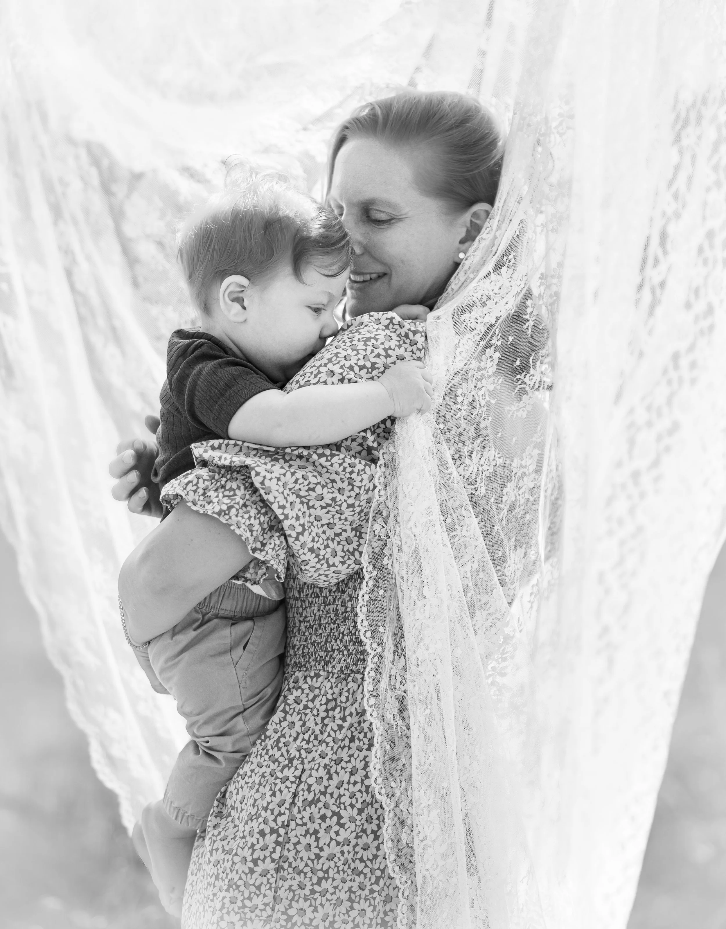 A woman and a young boy embrace and smile, with their faces close together, framed by a sheer white curtain.