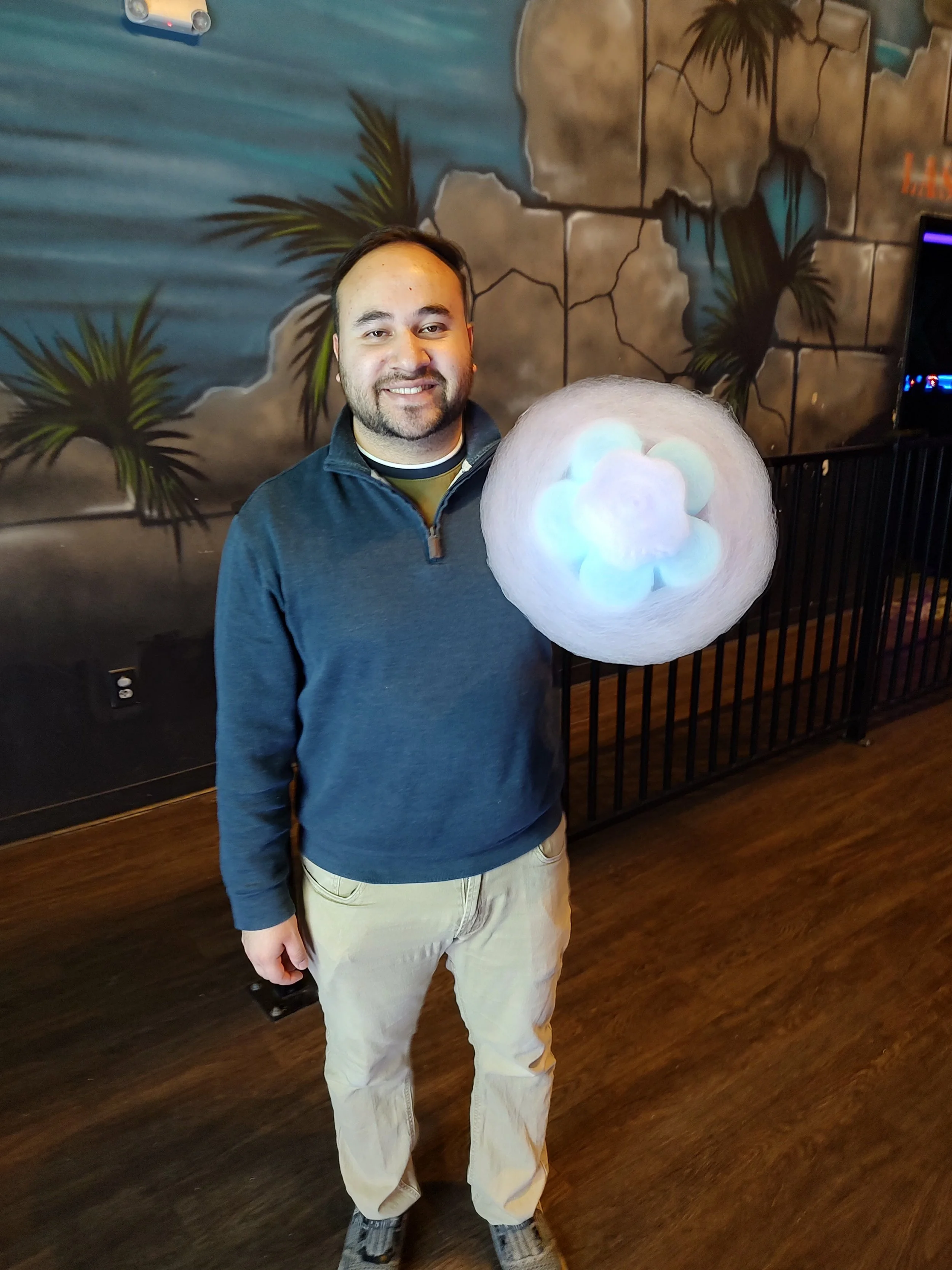 A smiling man holding a glowing cotton candy in an indoor arcade or entertainment venue with a tropical-themed mural in the background.