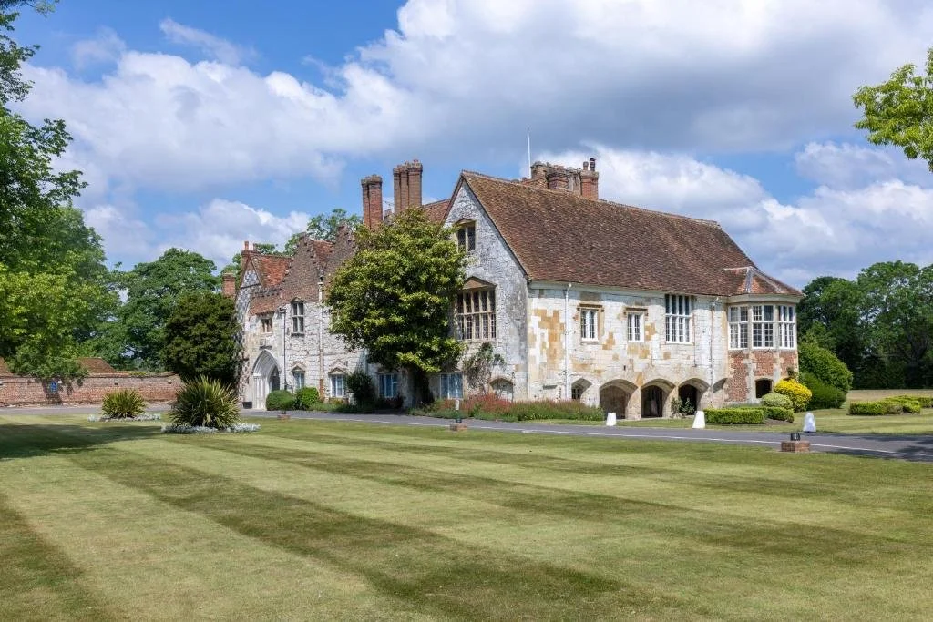 An old, large stone house with a red-tiled roof, multiple chimneys, and arched openings underneath, surrounded by well-kept green lawns and trees. A partly cloudy sky is visible above.