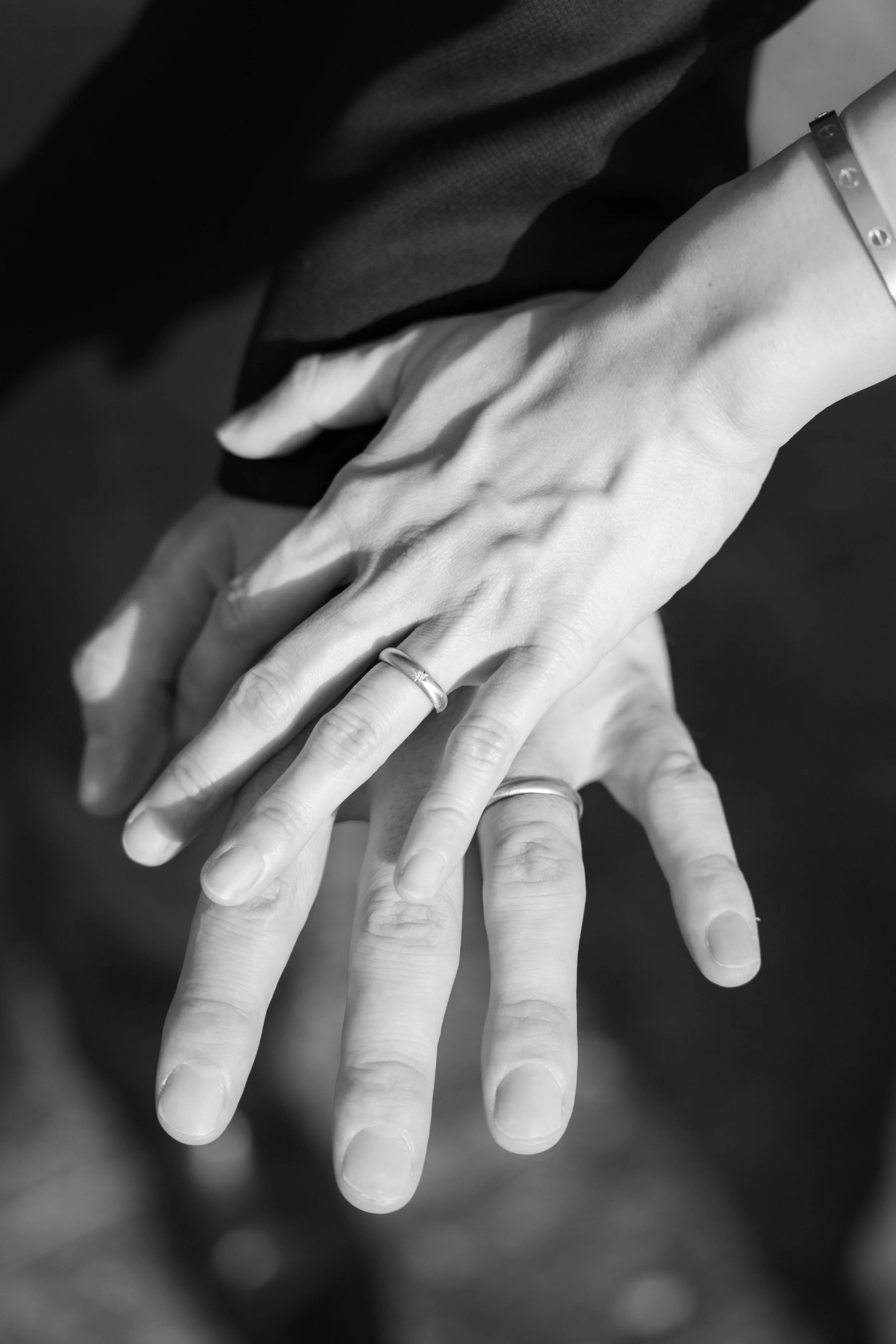 Close-up of two hands with wedding rings, one placed on top of the other, against a blurred dark background.