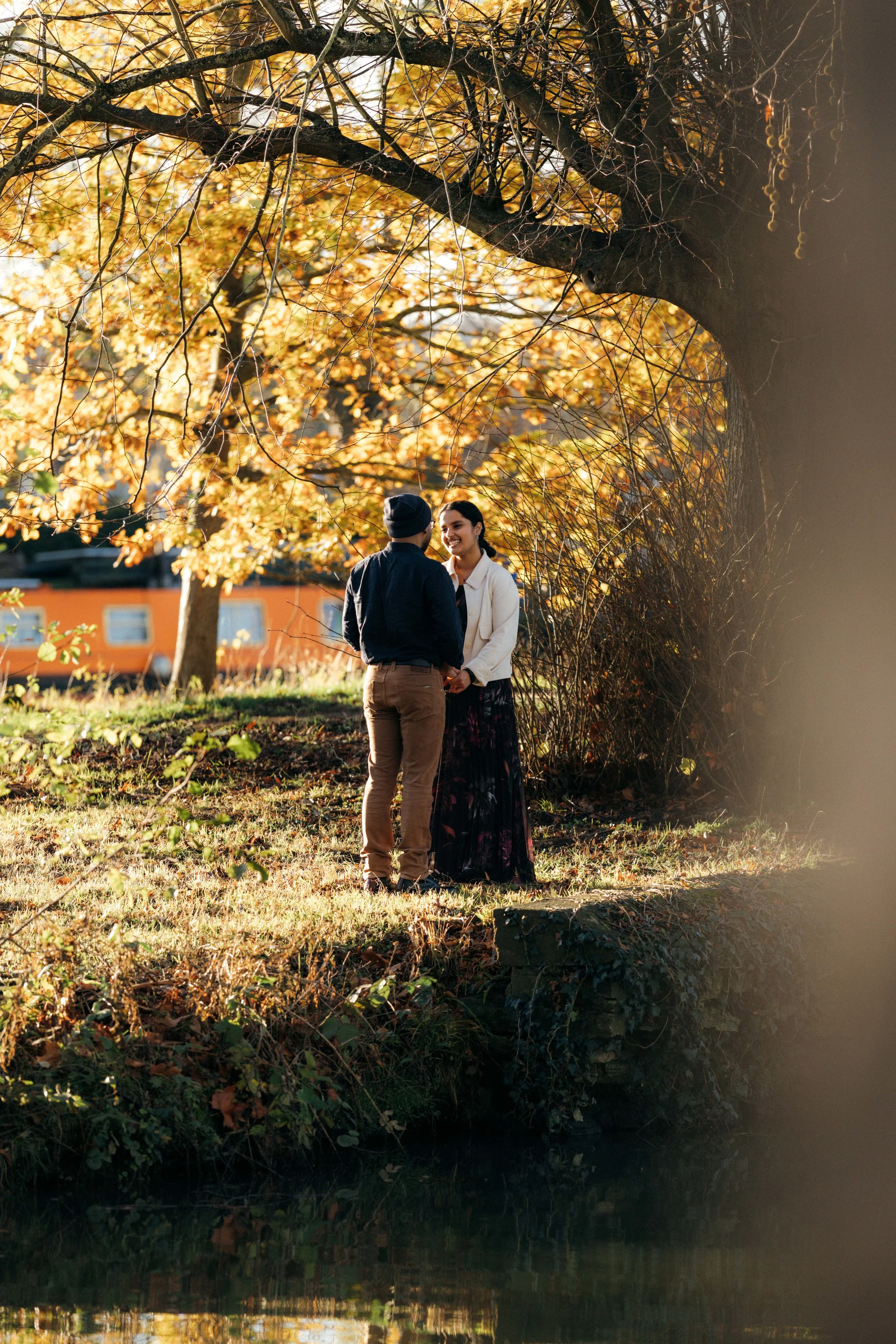 A couple standing on a grassy area near a tree beside a body of water, holding hands and smiling at each other. The scene is bathed in warm, golden sunlight with autumn leaves in the background.