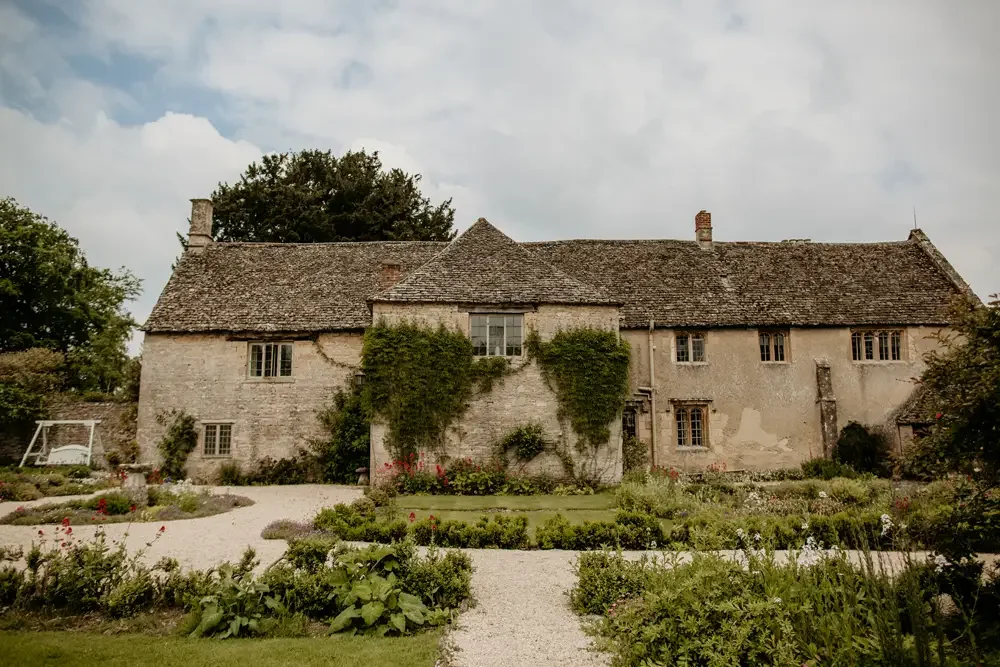 An old stone house with a weathered exterior, surrounded by a well-maintained garden with various plants and flowers, and a gravel pathway leading up to it.