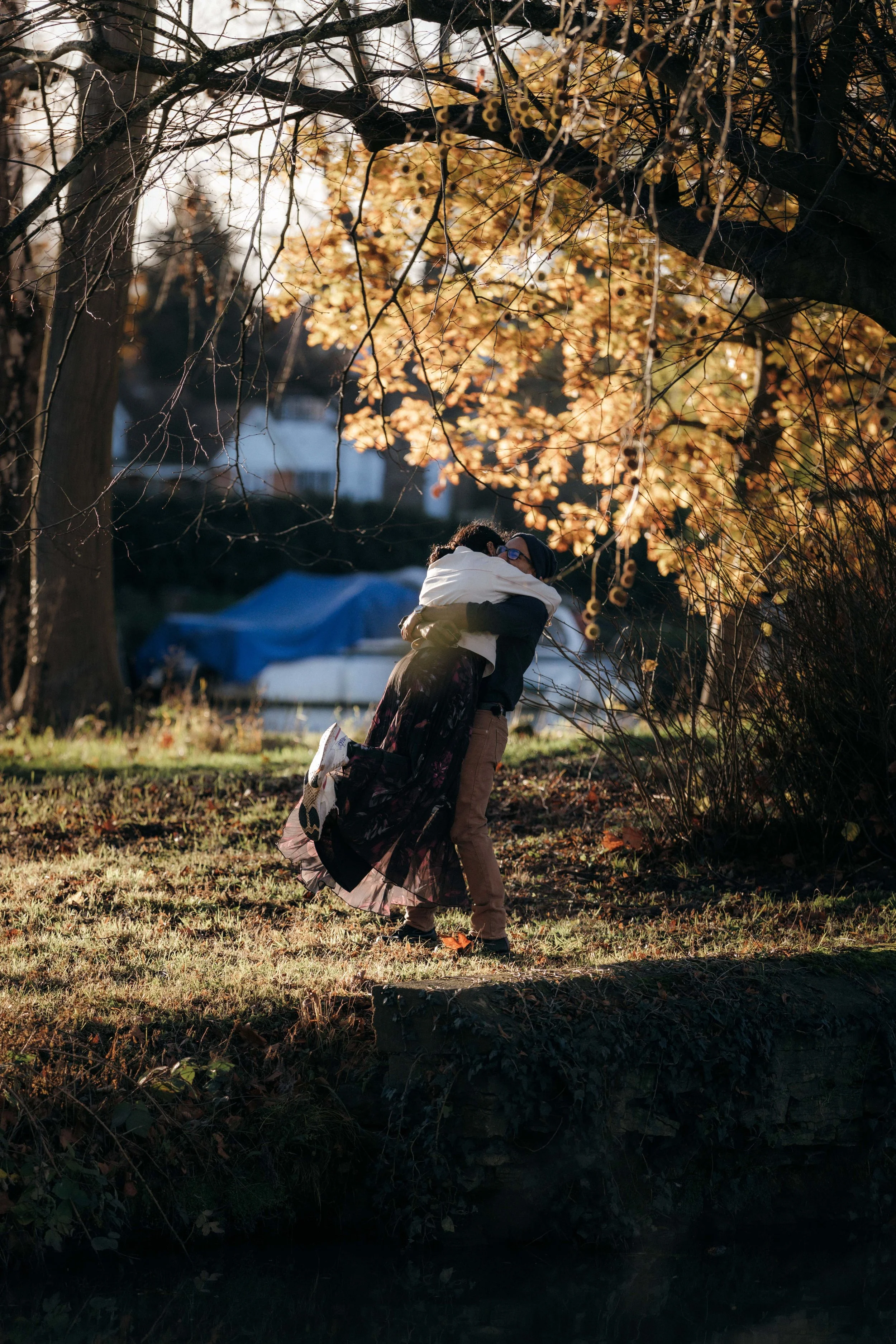 Two people hugging outdoors on a fall day, surrounded by trees with golden leaves.