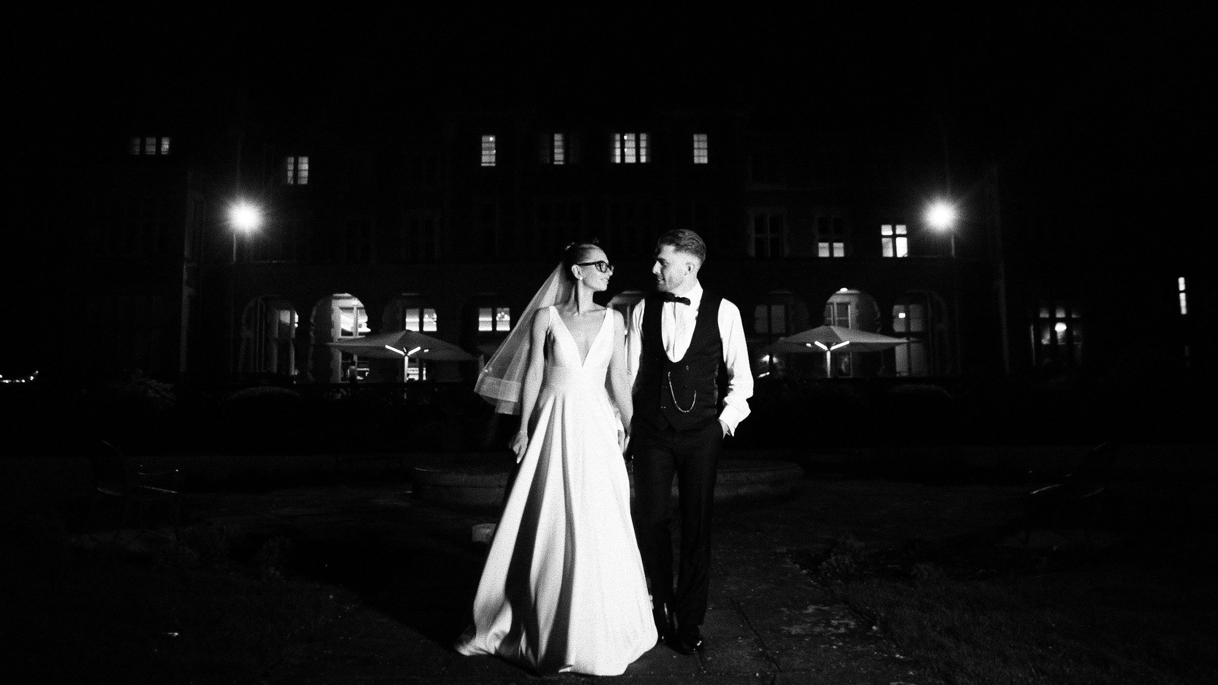 Black and white photo of a bride and groom walking together outdoors at night, holding hands, with a large building with illuminated windows behind them.