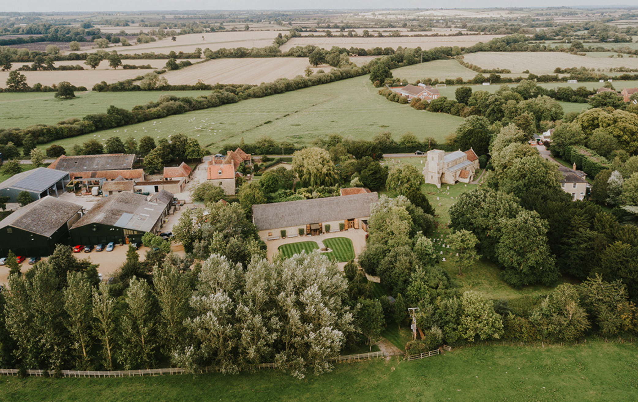 Aerial view of a rural landscape with green fields, trees, and farm buildings, including a large house with a well-maintained lawn and a church visible in the background.