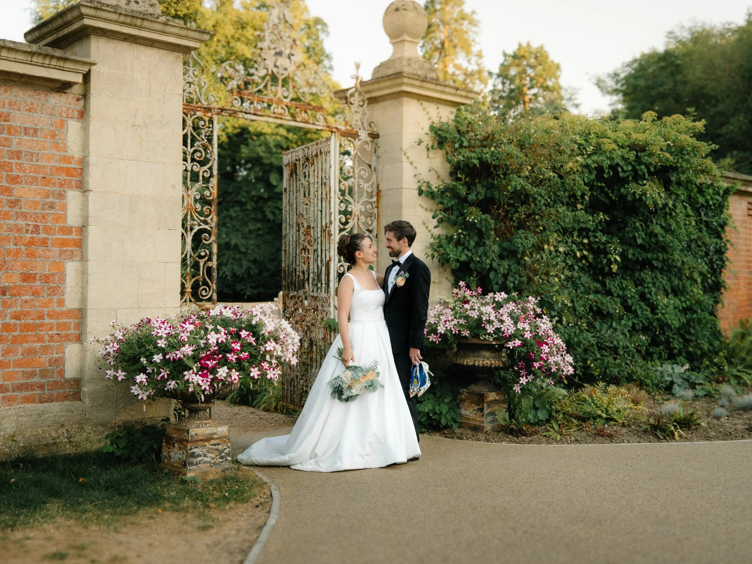 A bride and groom standing and smiling at each other near a decorative wrought-iron gate surrounded by greenery and pink flowers.