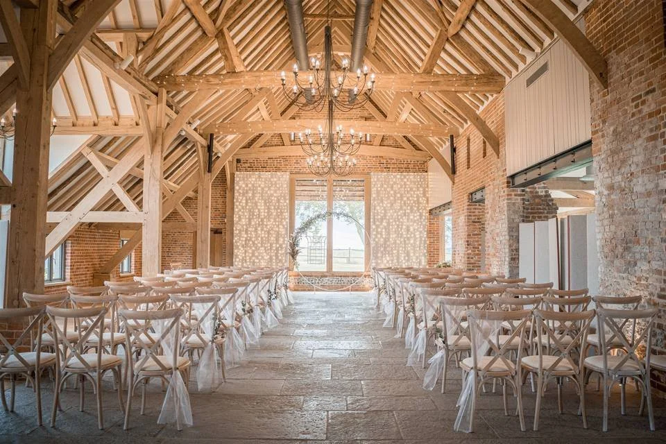 Interior of a rustic venue, Rackleys in Lane End, decorated for a wedding with rows of white chairs tied with white fabric, a wooden arch with flowers and greenery, and chandeliers hanging from a high, vaulted wooden ceiling.