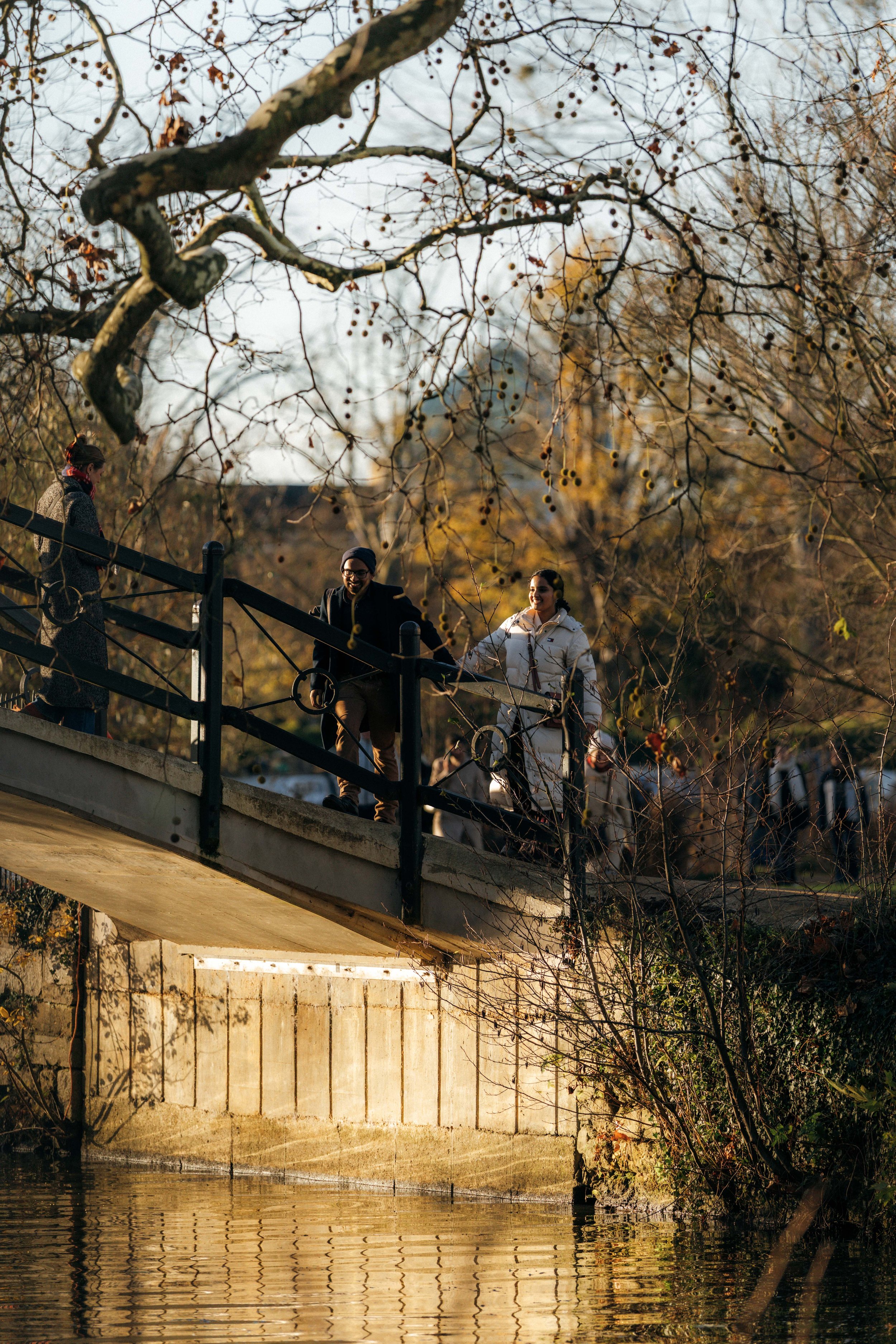 Three people walking on a small bridge over a waterway during autumn, with bare trees and sunlight in the background. Pre Proposal