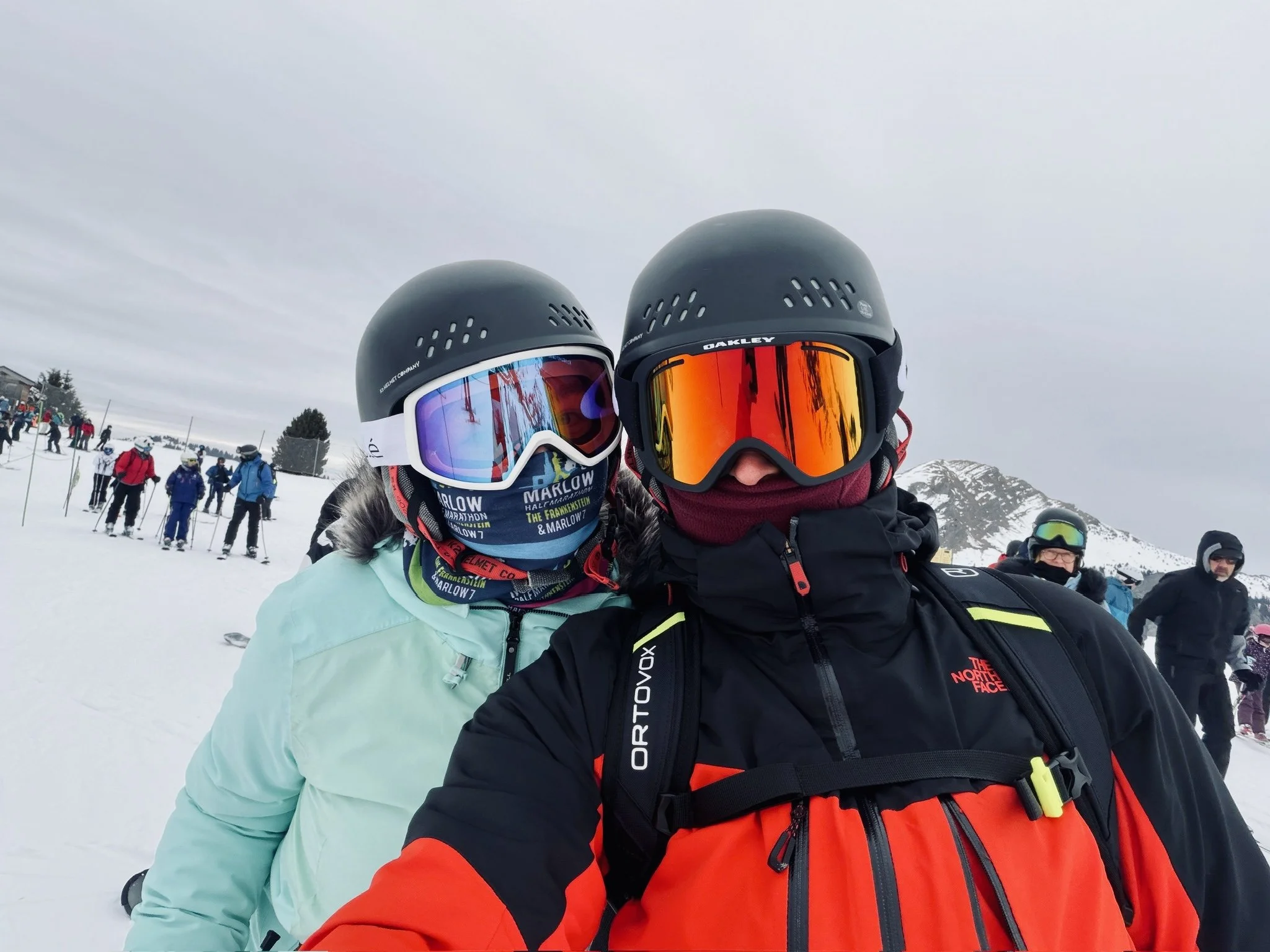 Two skiers in helmets and goggles taking a selfie on a snowy mountain, with a group of other skiers and snow-covered peaks in the background.
