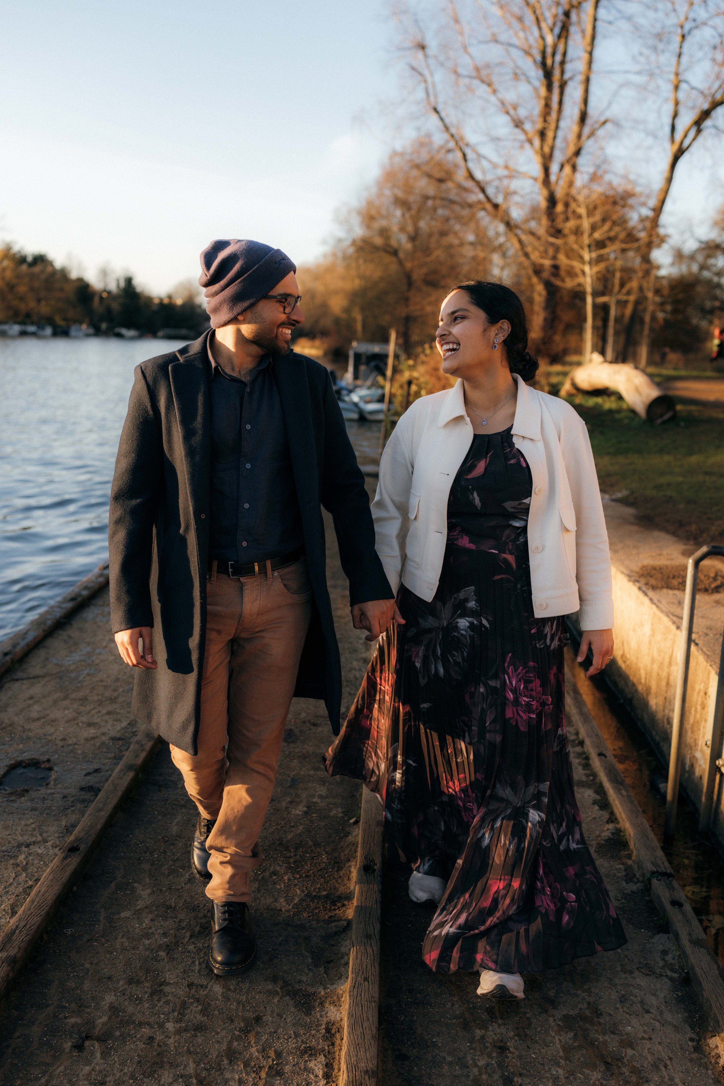 A man and woman walking hand-in-hand along a lakeside path, smiling and looking at each other during sunset, with leafless trees and boats in the background.