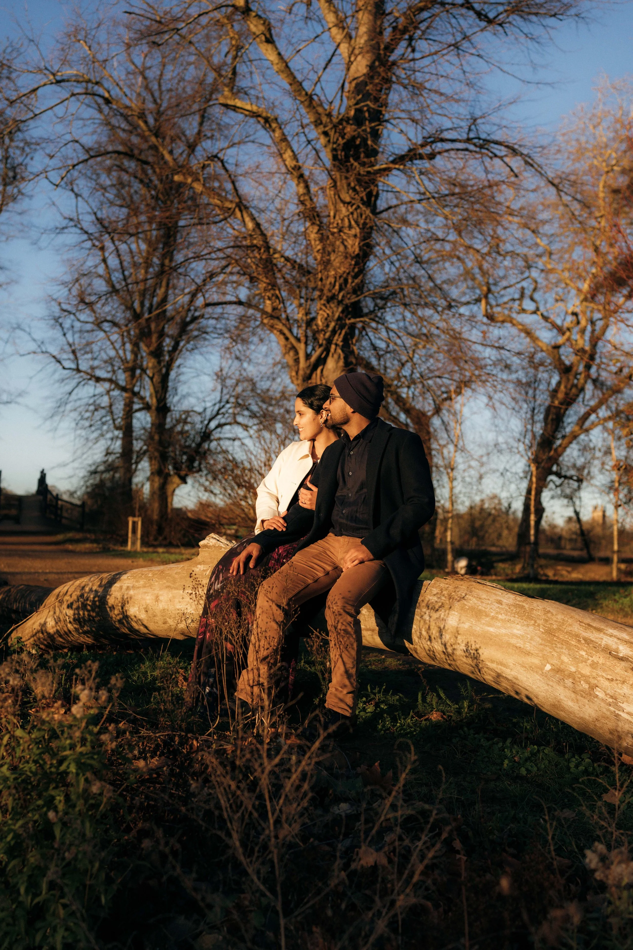 A man and woman sit on a fallen tree in a park during sunset, with bare trees in the background.