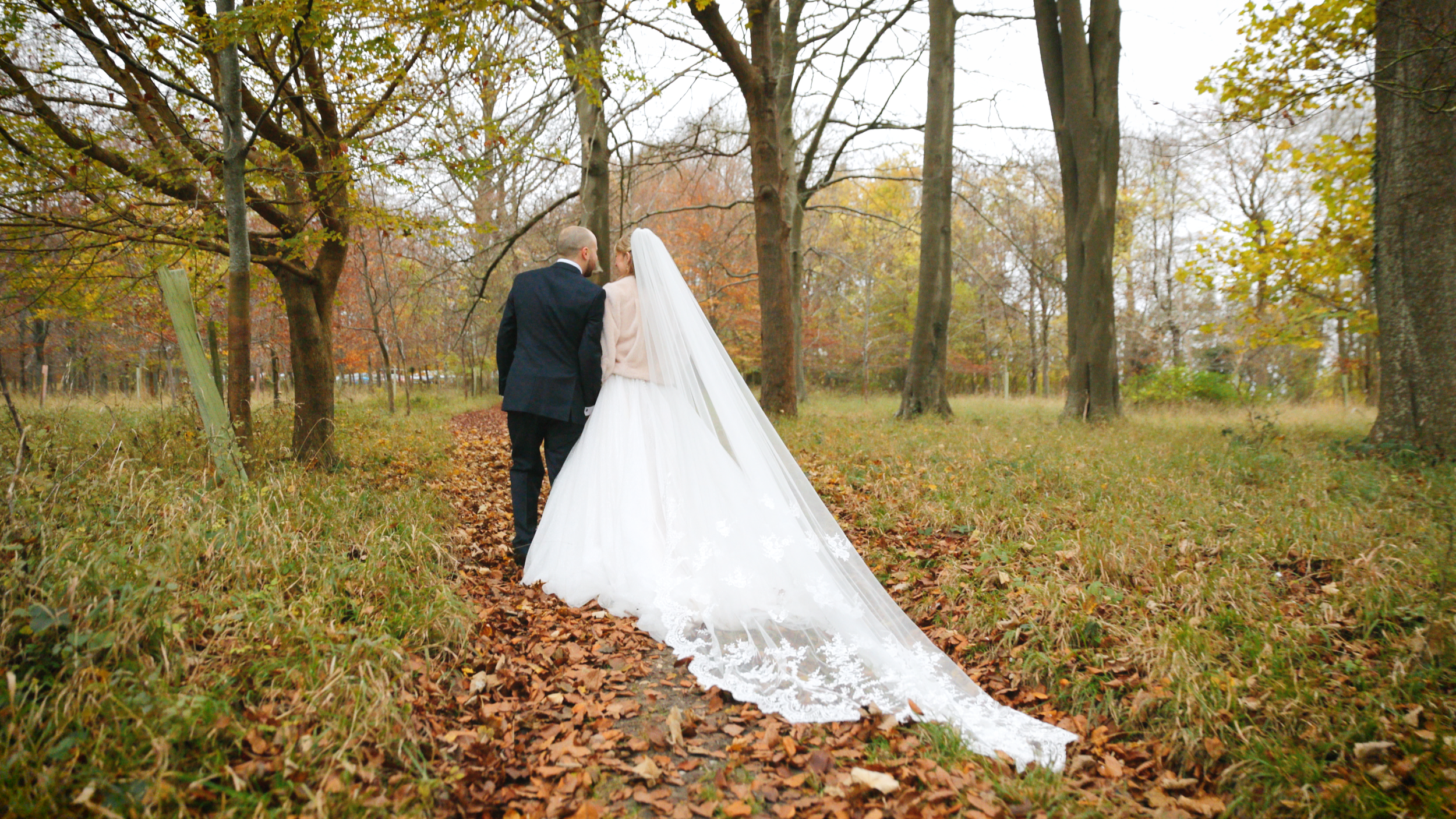 A bride and groom walking hand in hand on a leaf-covered trail in a wooded area during autumn. At Old Luxters Barn, Henley.