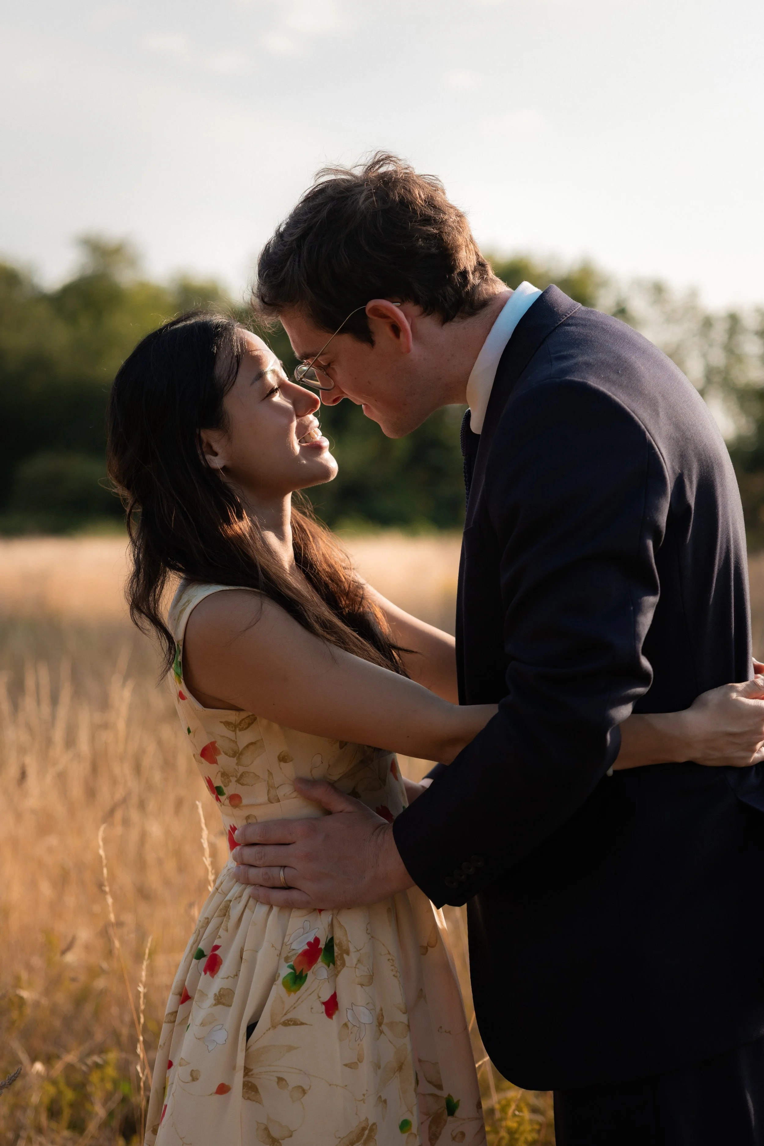 A man and woman in formal attire embrace and touch foreheads in an outdoor field during sunset, smiling at each other.