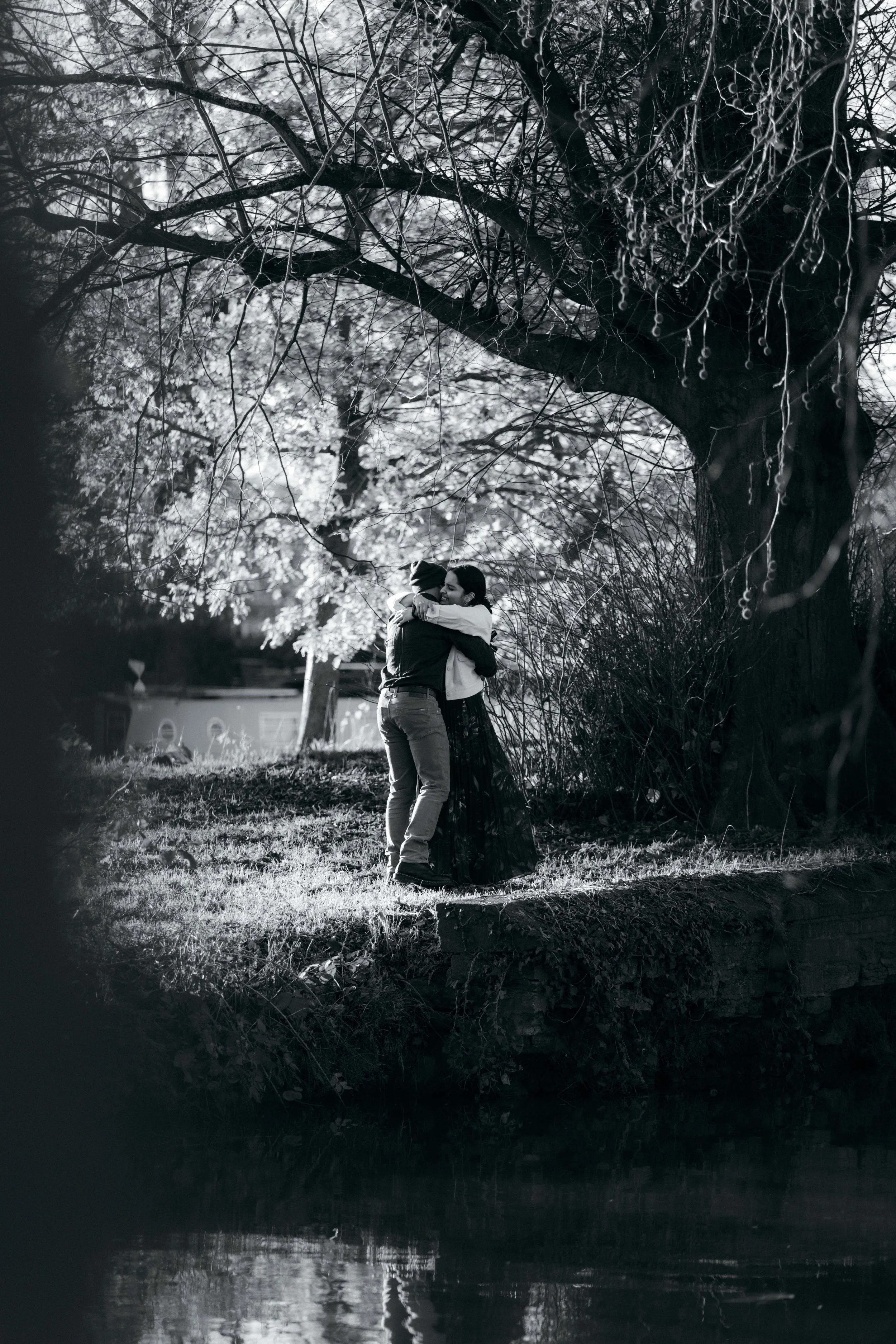 A black-and-white photo of two people hugging under a large tree next to a body of water, with the sunlight filtering through the branches.