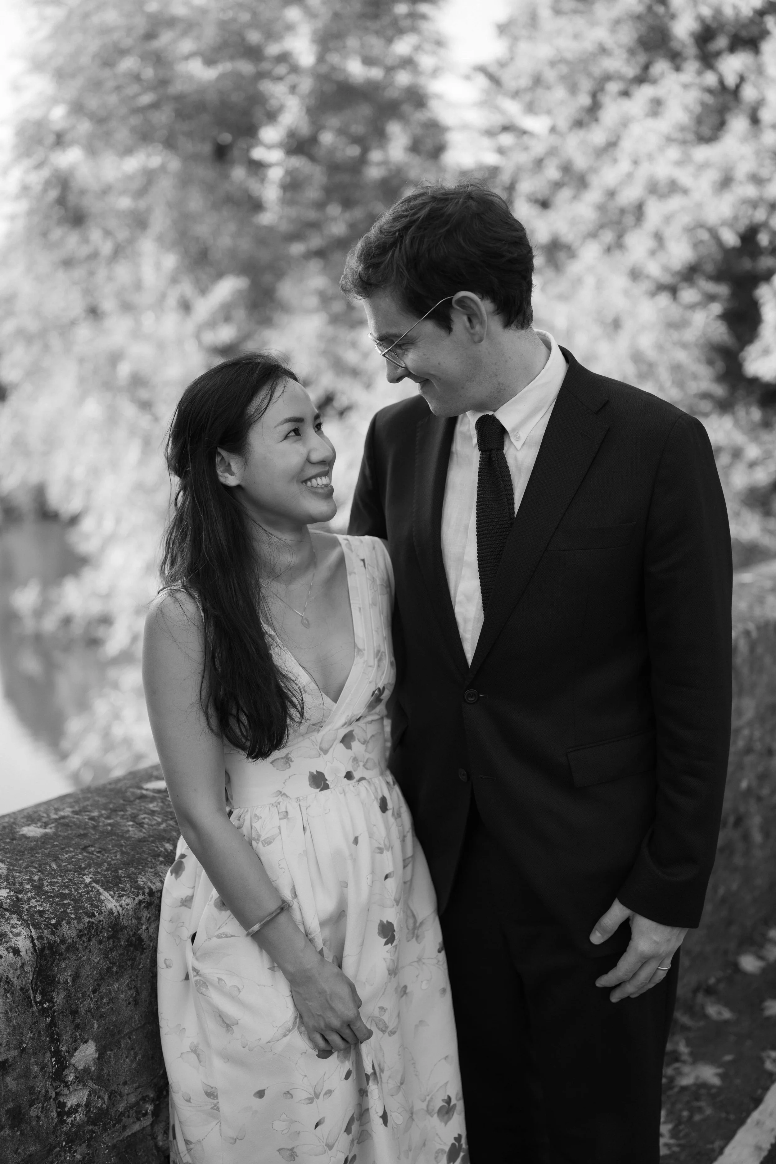 A woman and a man face each other smiling outdoors, with trees in the background. The woman is wearing a floral dress and the man is in a suit.