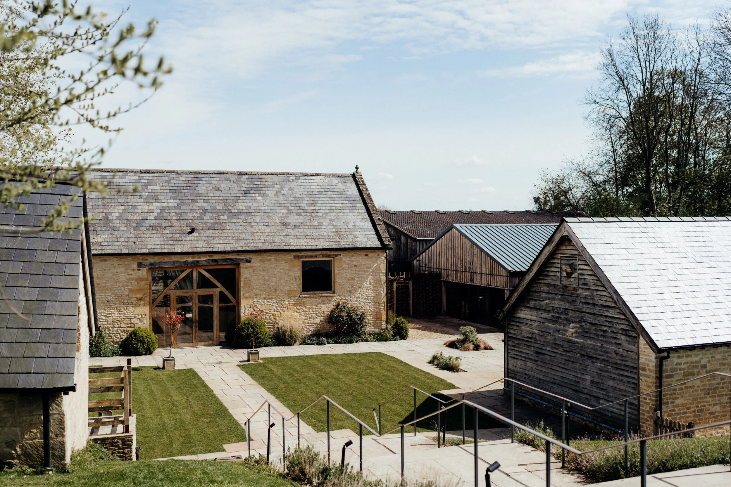 A courtyard with a well-maintained lawn, stone pathway, in front of rustic stone and wooden buildings under a partly cloudy sky, with trees in the background.