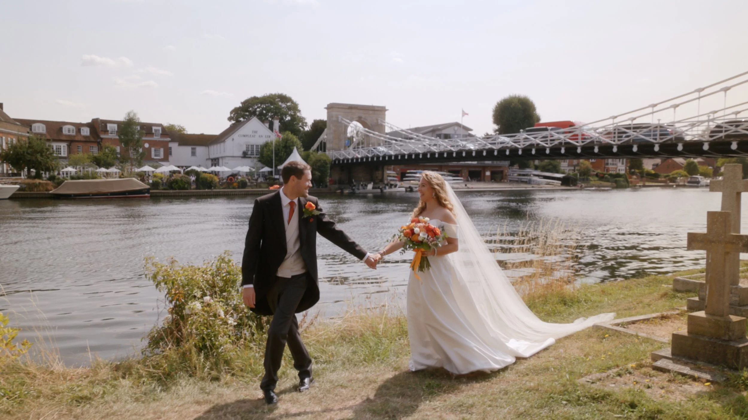 A bride and groom holding hands near a river, with a bridge and riverside buildings in the background.