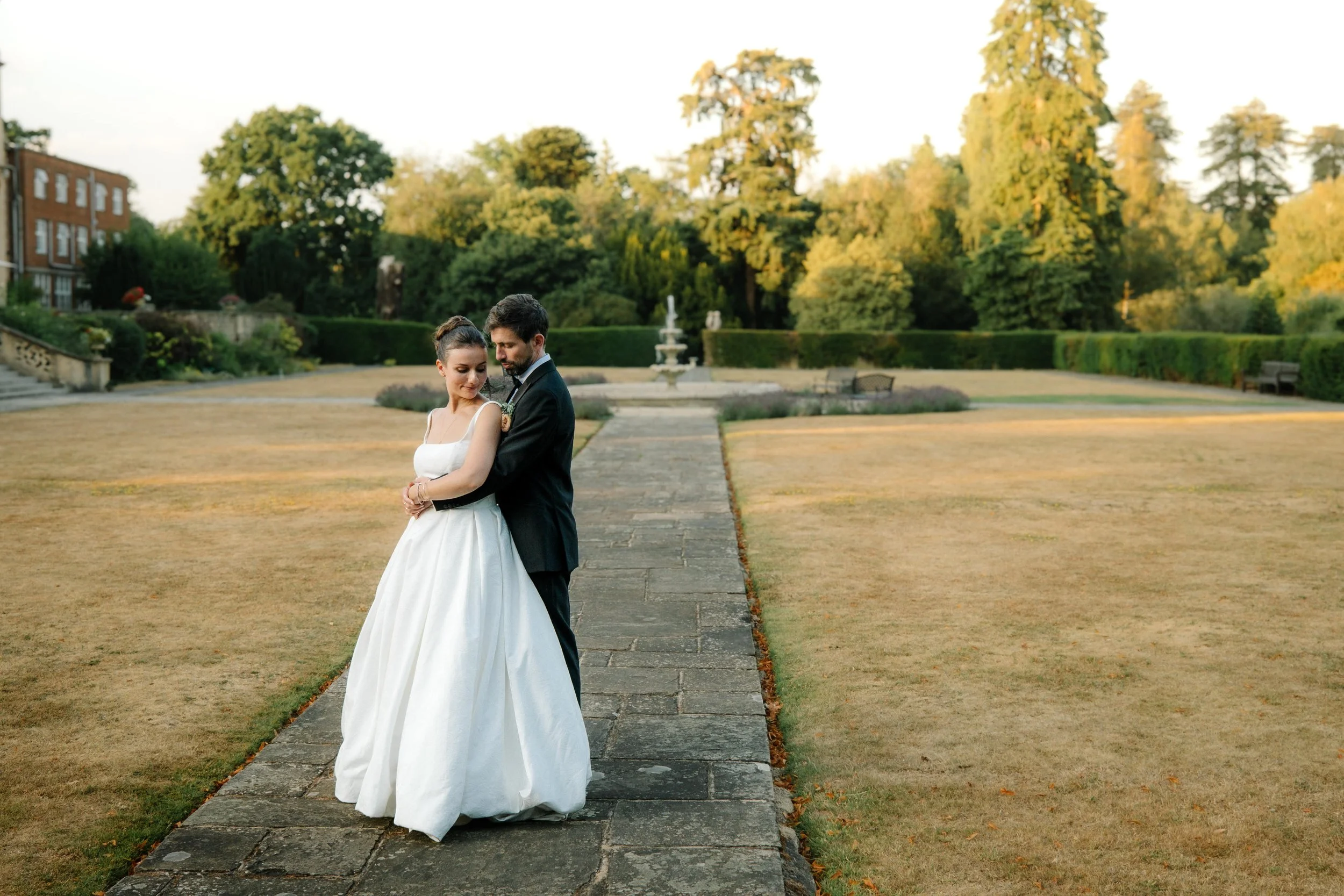 Bride and groom standing together on a stone path in a park at east hampstead park, wokingham, surrounded by trees and greenery, during sunset.