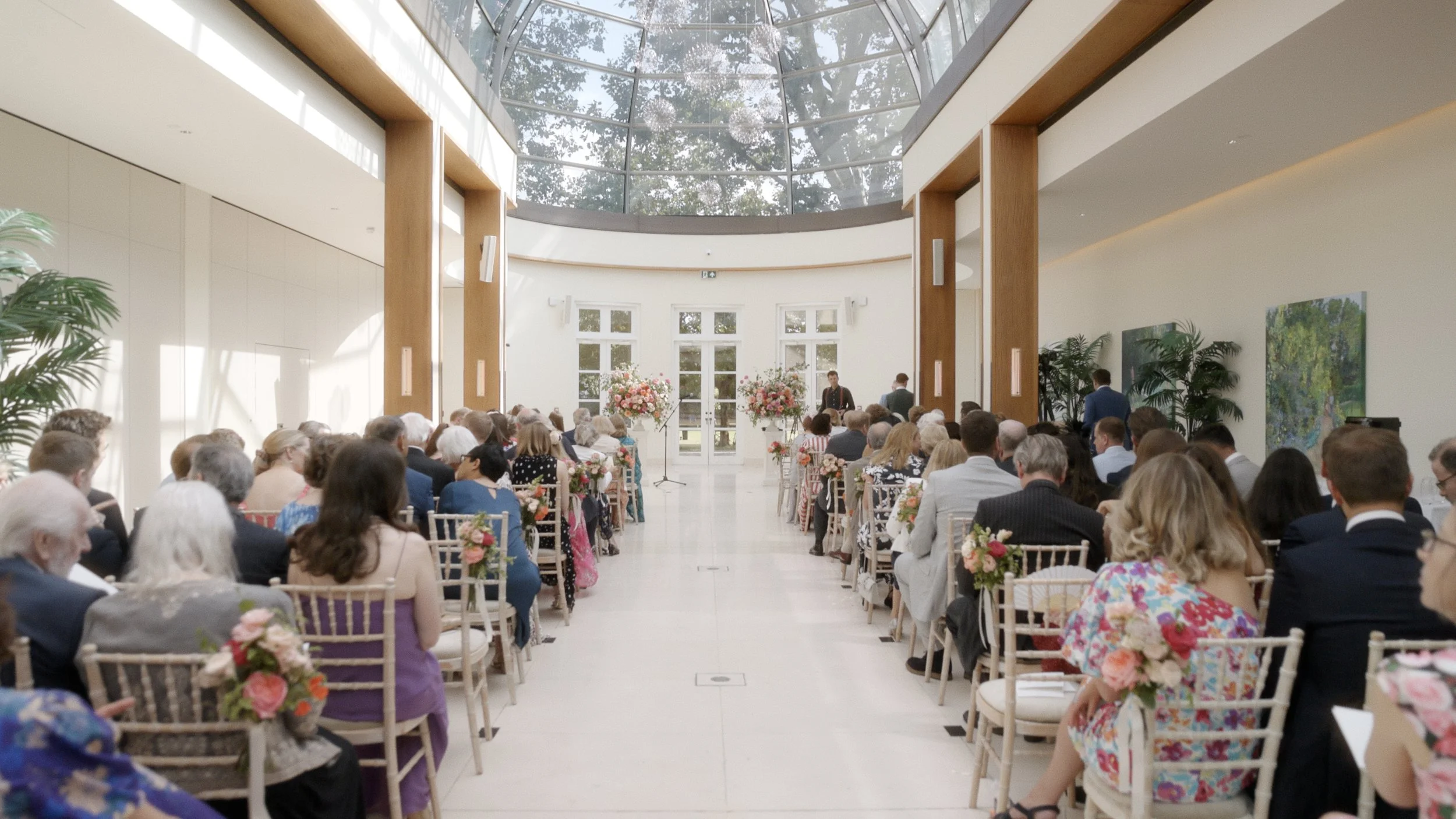 Indoor wedding ceremony with guests seated on either side of the aisle, floral arrangements, large windows, and a glass ceiling.