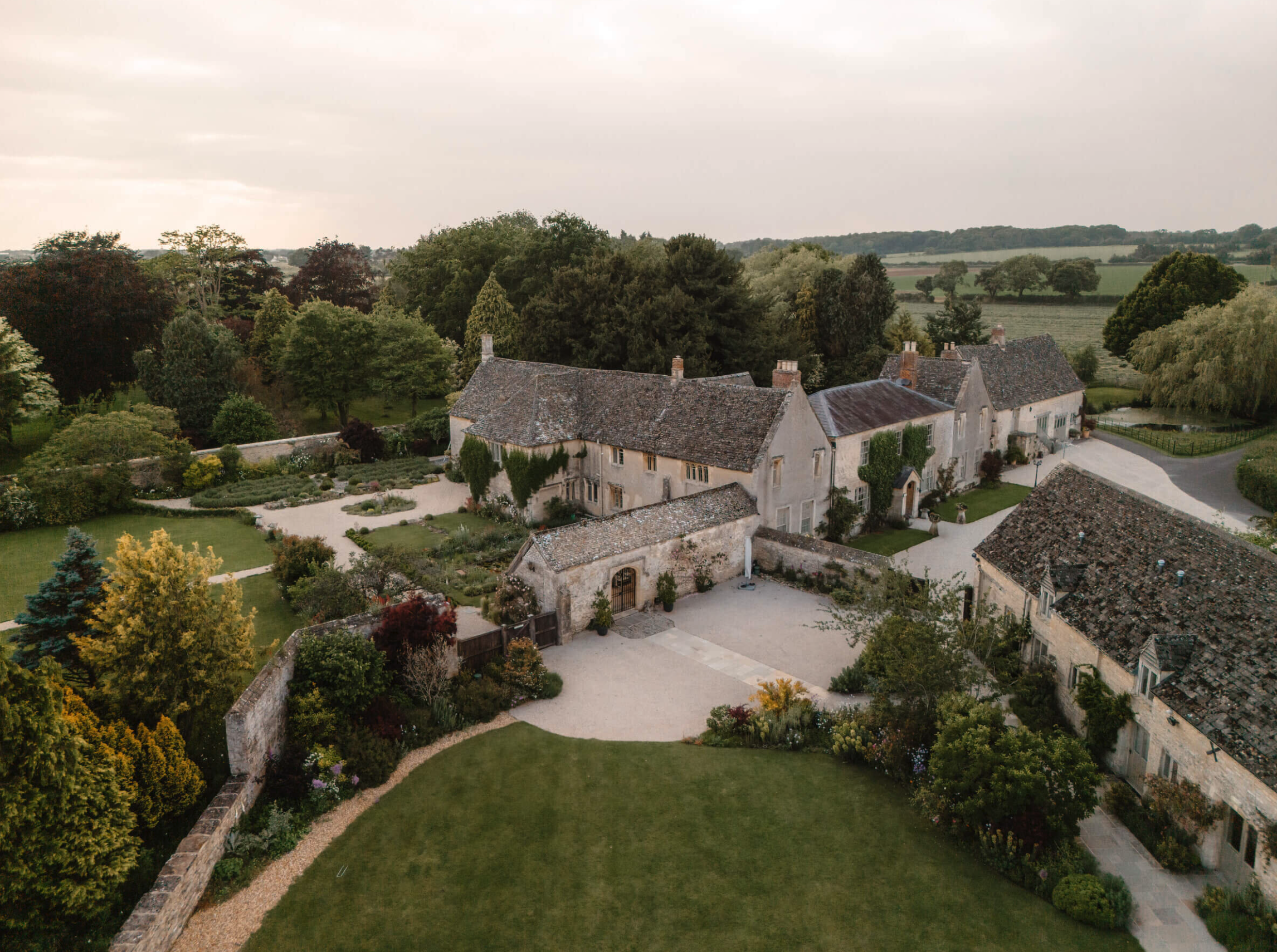 Aerial view of a historic stone estate with multiple buildings, surrounded by well-maintained gardens and lush trees, amid a rural landscape with open fields and a cloudy sky.