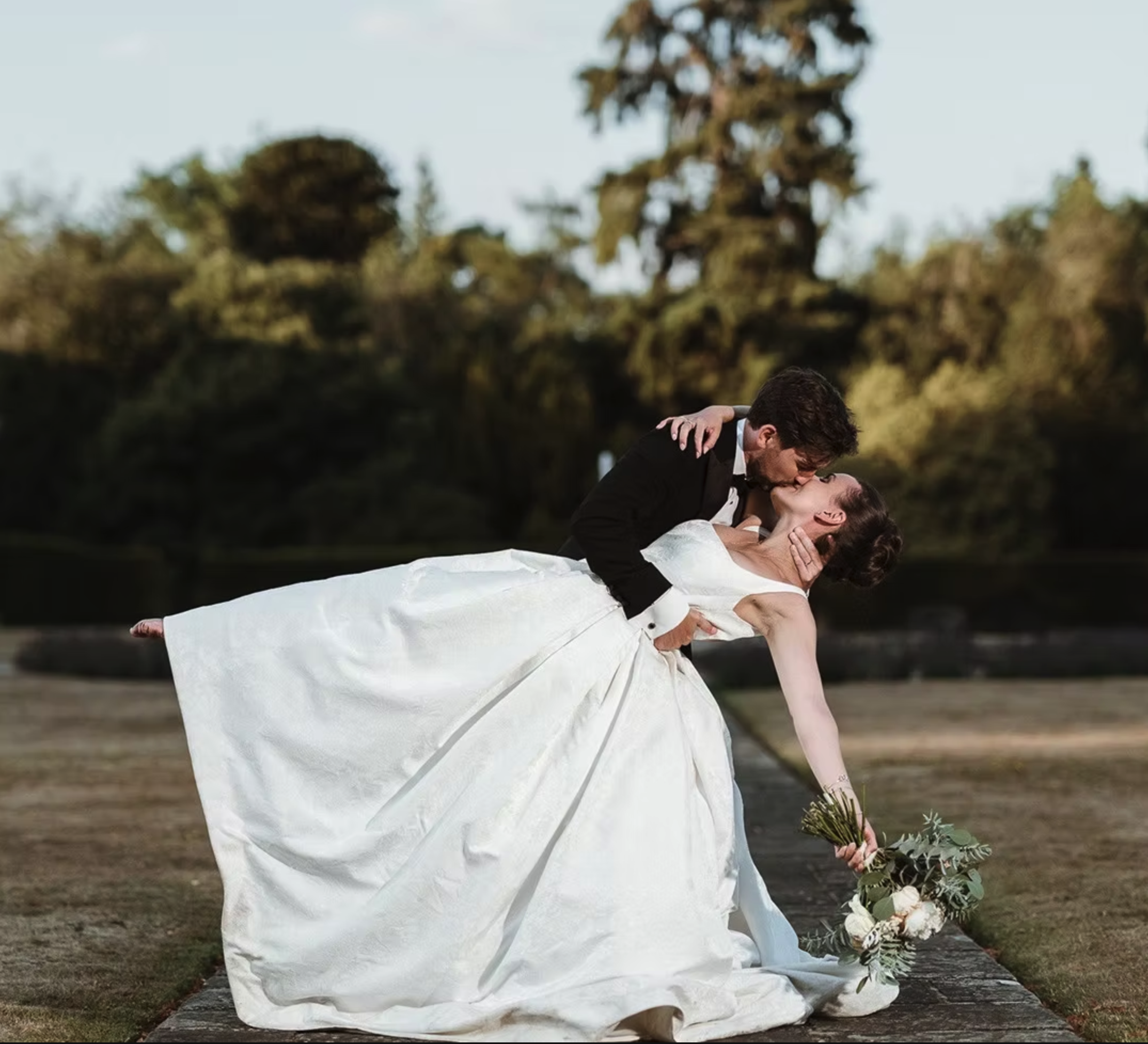 A bride and groom share a kiss outdoors, with the bride in a white wedding dress and the groom in a black tuxedo. The bride dips and extends her hand holding a bouquet of flowers, and greenery, towards the ground, while the groom holds her securely.