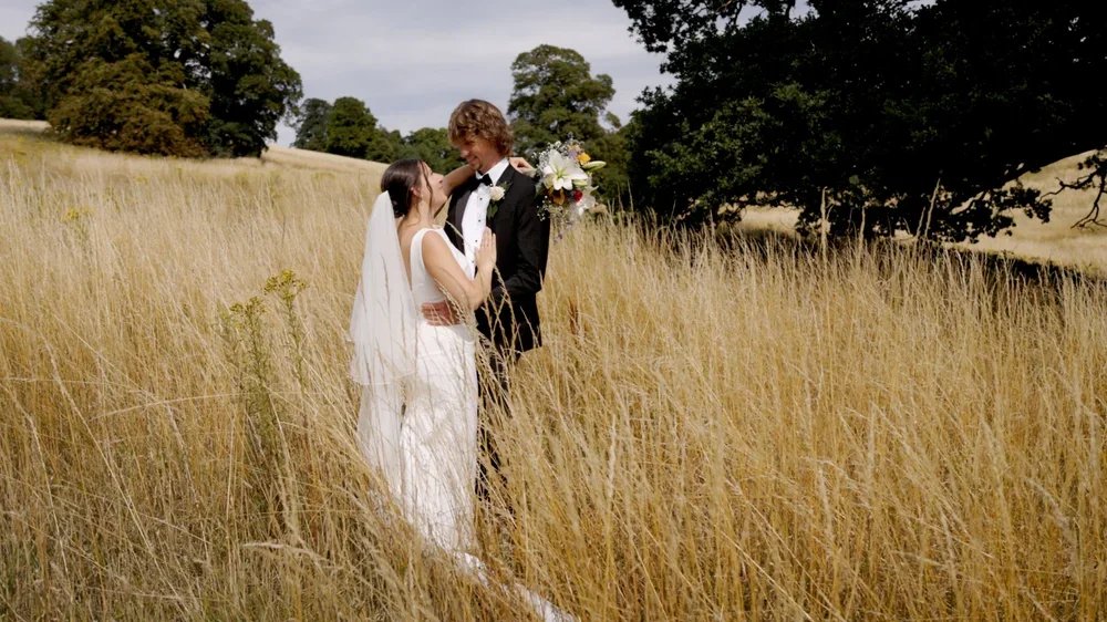 A bride and groom standing close together in a field of tall, golden grass with trees in the background, during a wedding photo shoot.