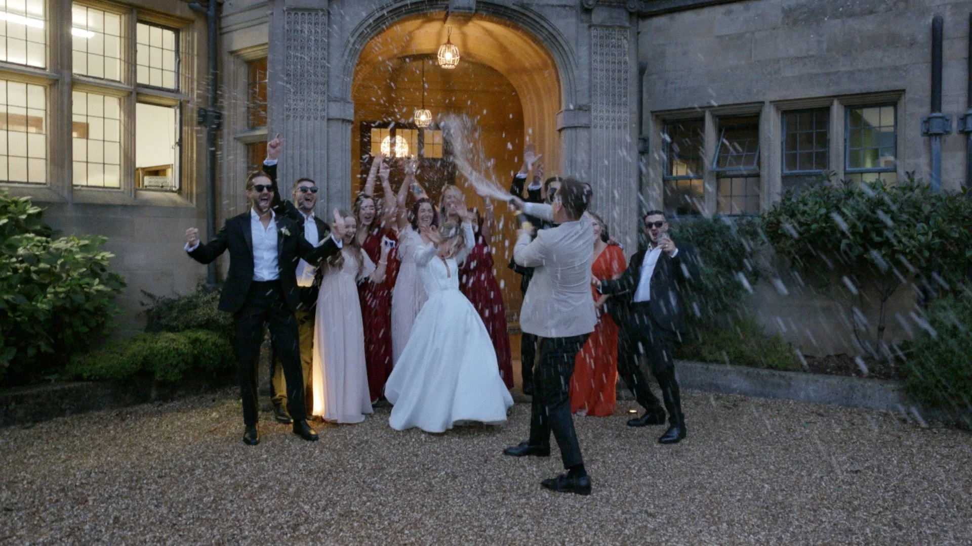 A wedding celebration with a group of people, including the bride in a white gown and groom, outside a stone building, being hit with a cake by the bride, while guests cheer they're at the Coombe Lodge, Blagdon