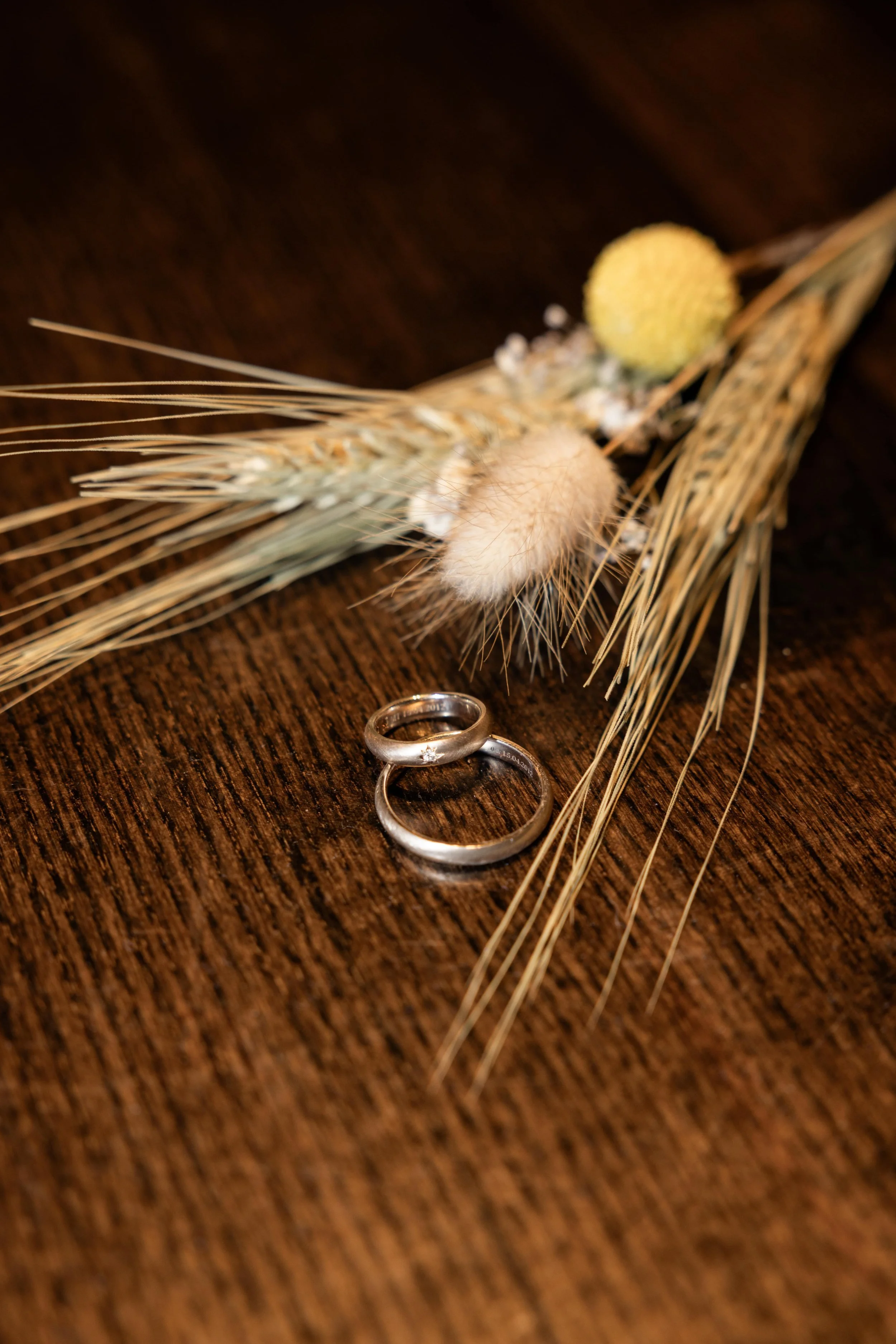 Two silver wedding rings on a wooden surface, with dried wheat and flowers in the background.