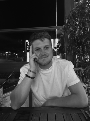 A young man (wedding videographer) with short hair and a beard sitting at a table in a restaurant or cafe, resting his head on his hand, smiling slightly, with a window and indoor plant in the background, in black and white.