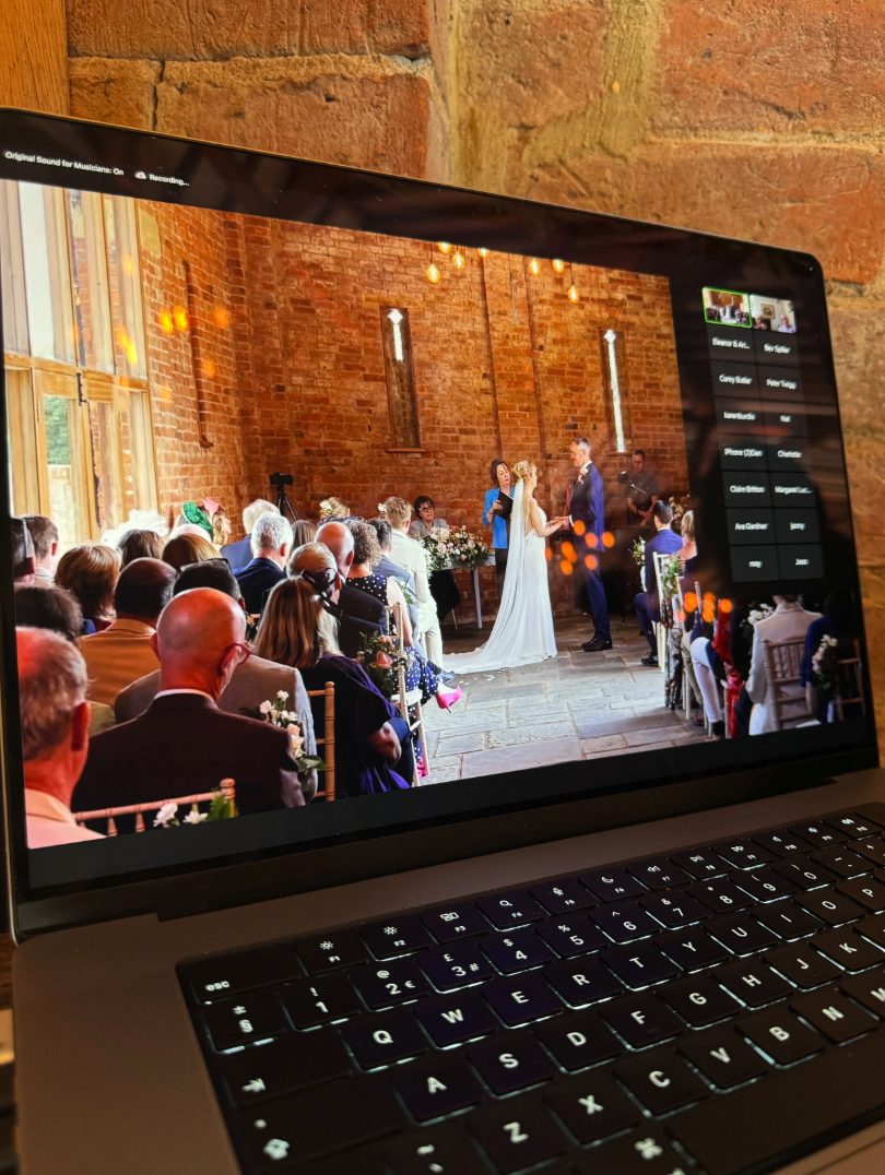 A laptop screen displaying a wedding ceremony live stream inside a church with brick walls and hanging lights, showing a bride and groom exchanging vows in front of seated guests.