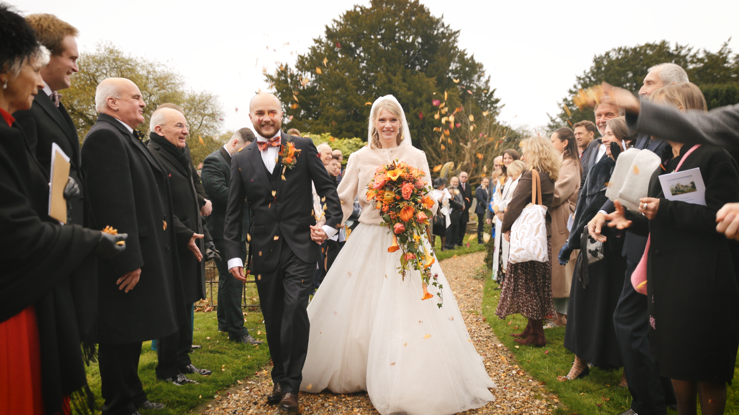 Bride and groom walking hand in hand down an outdoor aisle while guests on both sides celebrate and throw confetti on a cloudy day.