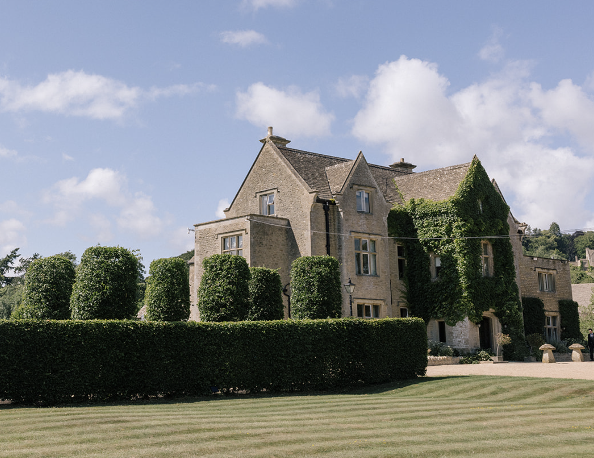 A large, historic stone house with a steeply pitched roof, surrounded by well-manicured hedges and trees, under a partly cloudy sky.