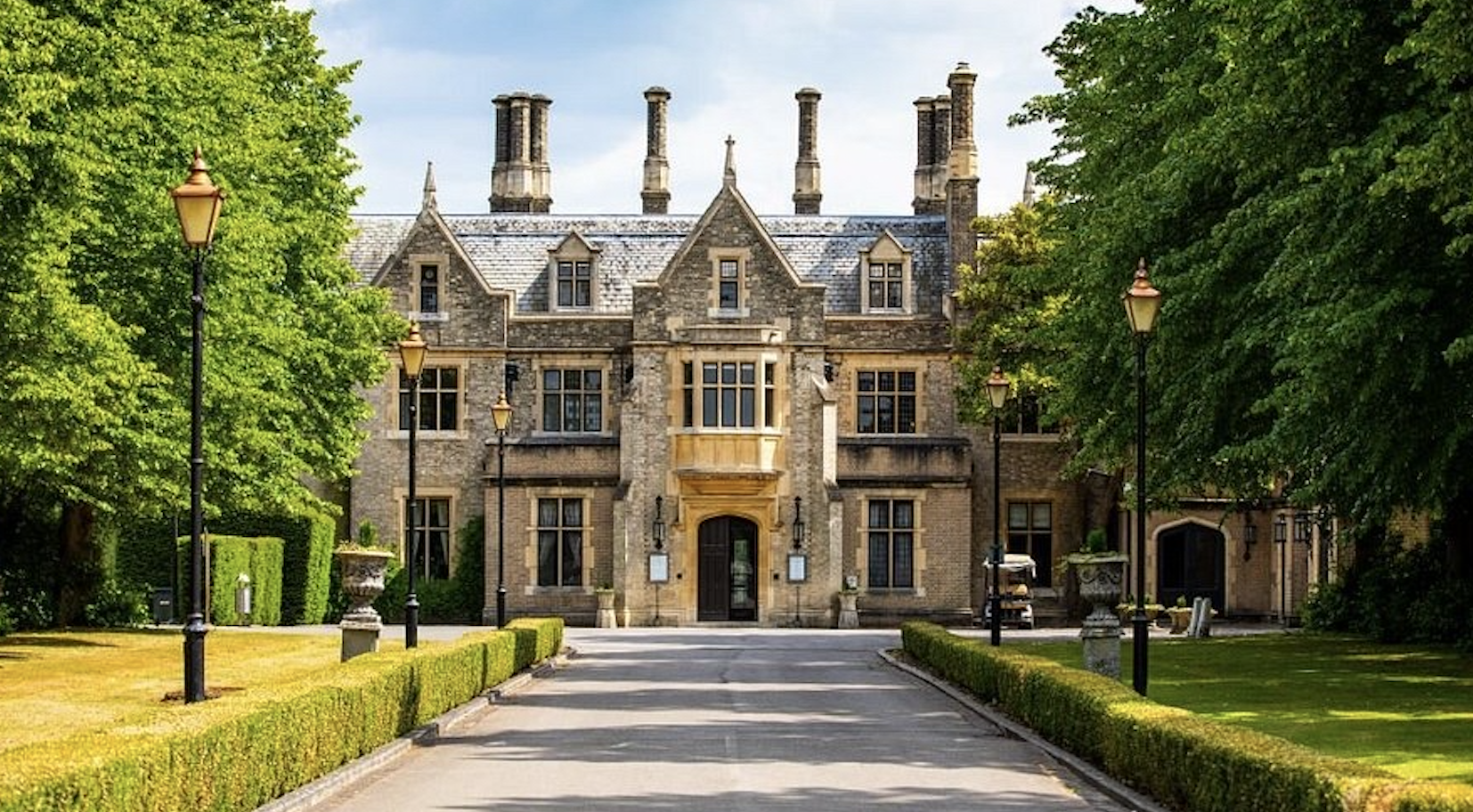 A large, historic stone mansion with multiple chimneys and gothic architectural features, surrounded by lush green trees and manicured lawns, with a paved driveway leading up to the entrance and vintage-style lamp posts along the path.