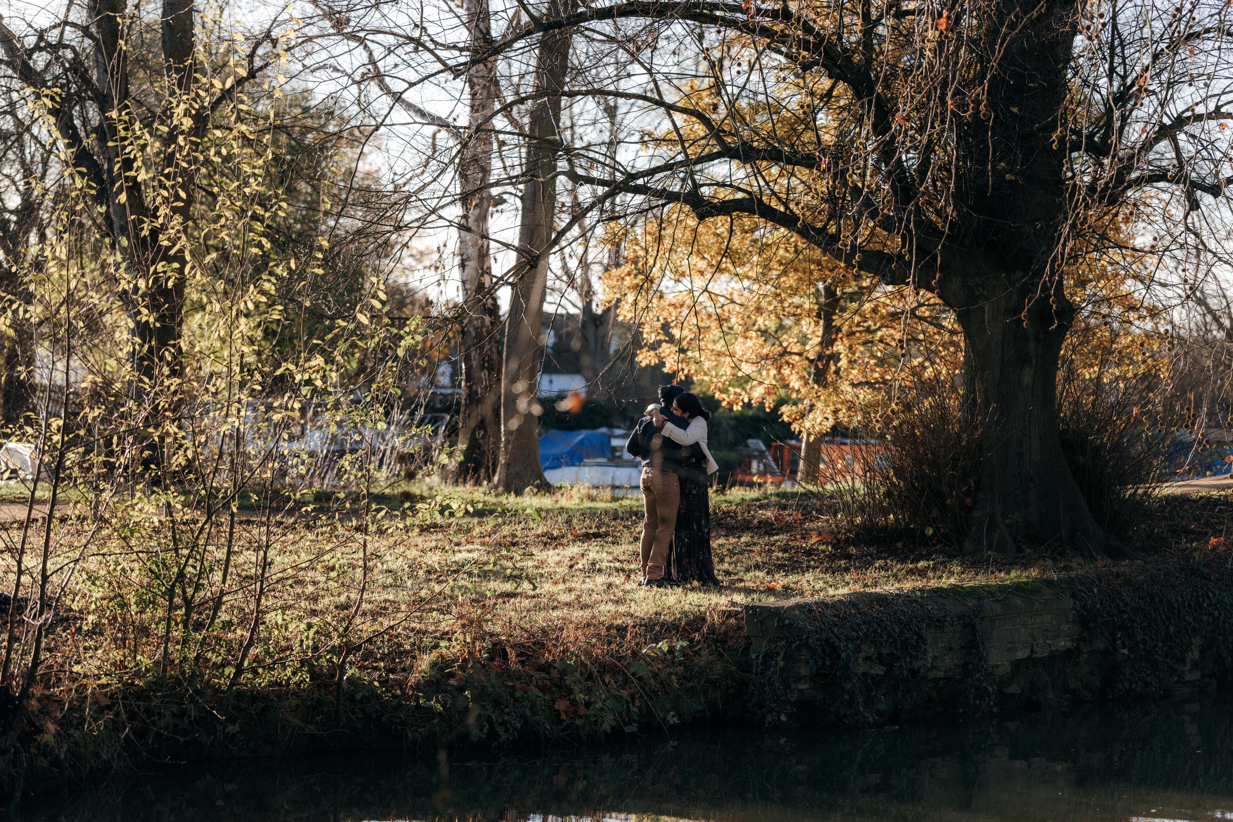 A couple is dancing or hugging outside under a large autumn tree with golden leaves, near a small waterway, during late afternoon sunlight.