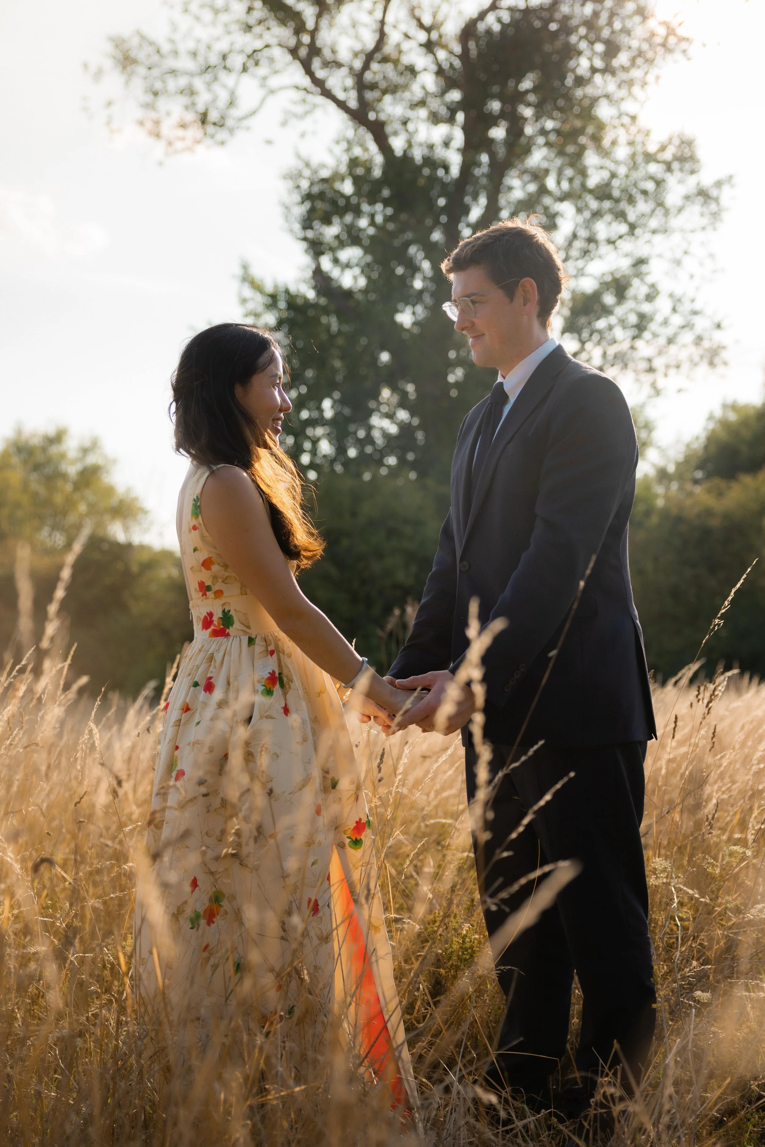 A man and woman stand in a field of tall grass, holding hands and looking at each other, with trees in the background and sunlight shining behind them.