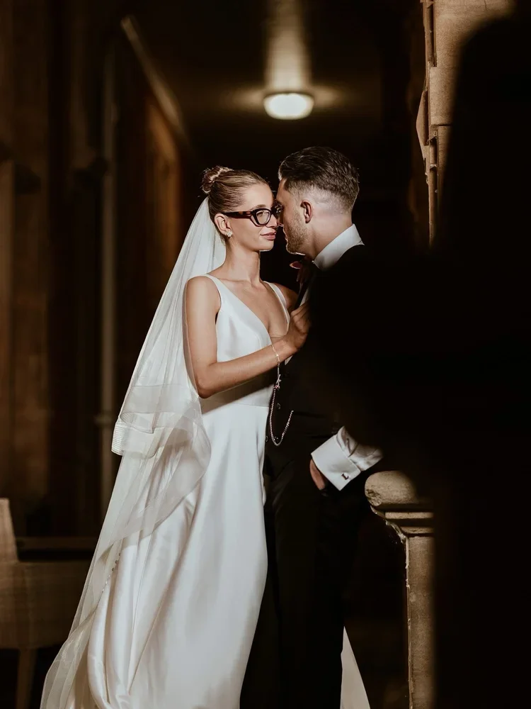 A bride and groom standing close together in a dimly lit hallway, with the bride wearing a white wedding dress and veil, and the groom in a black tuxedo. They are gazing into each other's eyes, creating an intimate moment. At East Hampstead Park