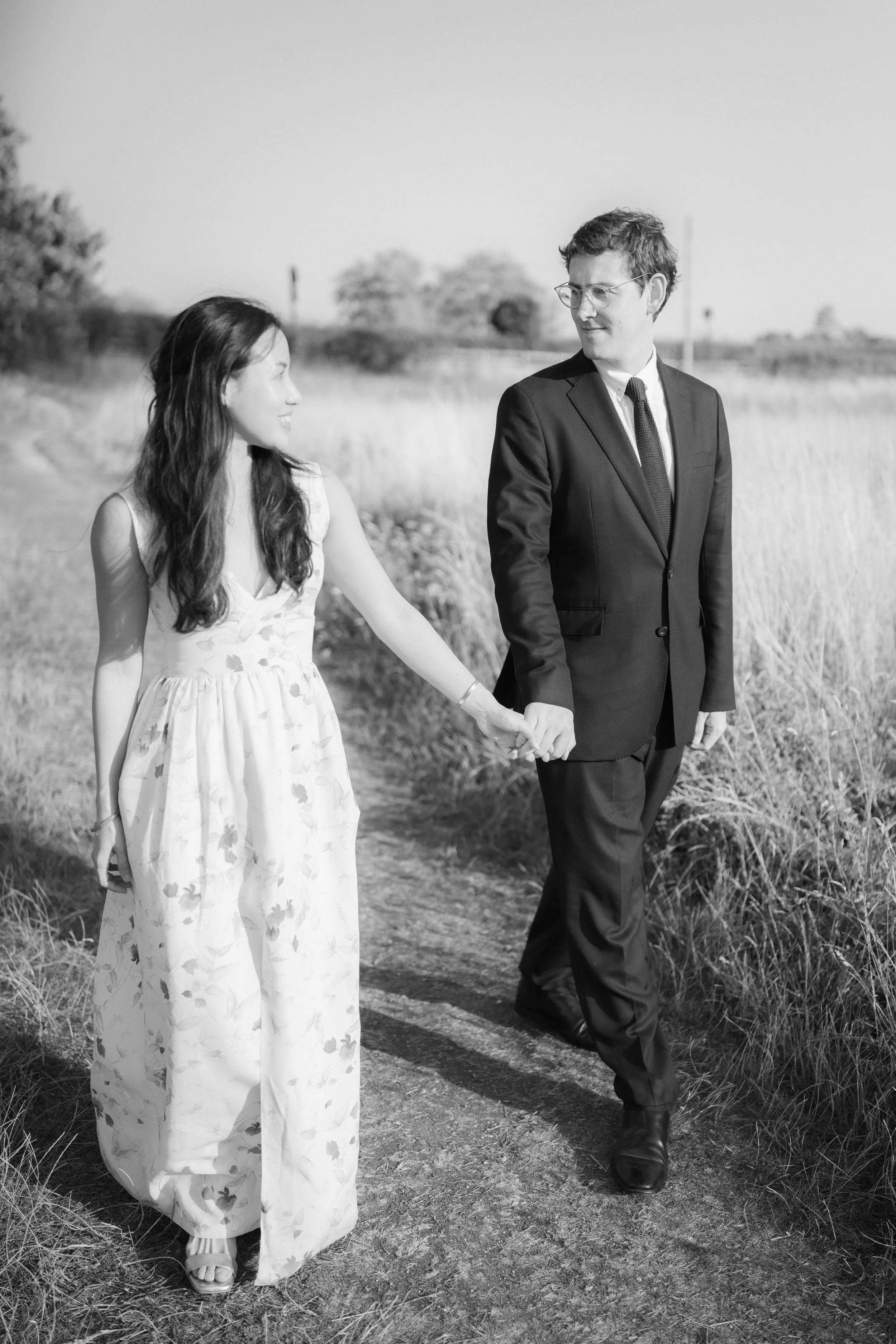 A couple walking hand-in-hand on a narrow dirt path in a grassy field. The woman is wearing a sleeveless floral dress, and the man is in a black suit with a tie and glasses. Both look at each other as they walk.